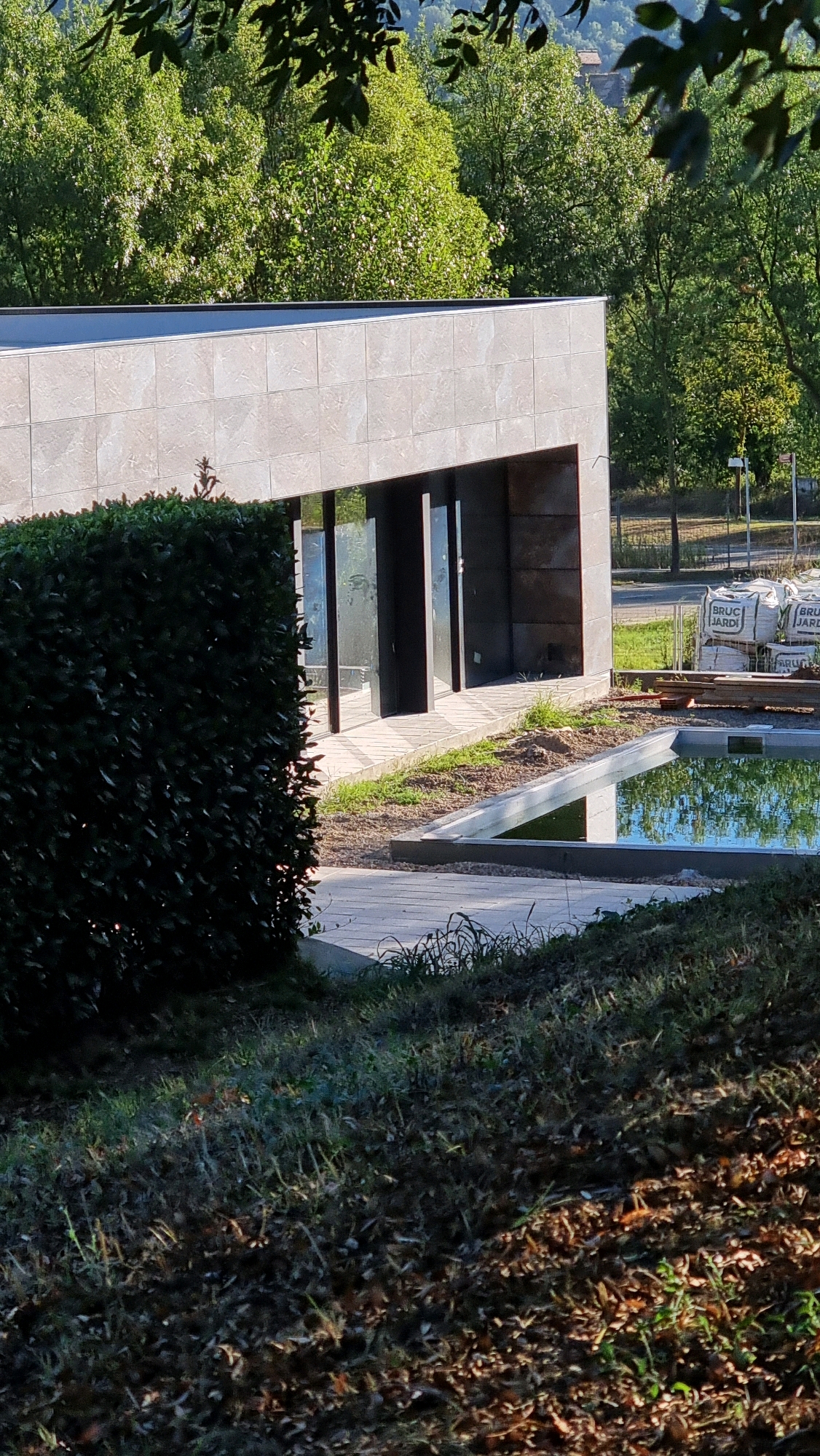 The image shows a modern building with large glass doors and windows, partially obscured by a dense green hedge. A shallow, rectangular pool of green water is visible in the foreground, bordered by concrete. The building's exterior is clad in large, light-colored tiles. In the background, a stack of white bags labeled "BRUGARDI" is present, along with wooden planks. The scene is set against a backdrop of lush green trees and foliage. There are no people visible in the image. The location is identified as Sant Joan les Fonts, Spain.