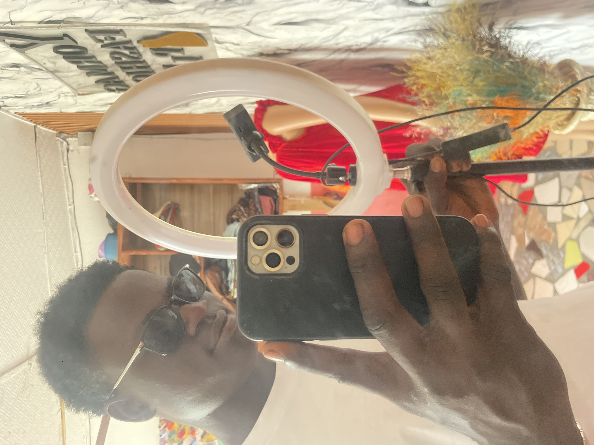 A person wearing sunglasses is holding a black iPhone in front of a ring light. The background includes items such as a mannequin head and decorative items attached to the wall. The image appears to have been taken in a room within a building, and, based on additional metadata (if available), the location is within the city of Badagry, Nigeria.