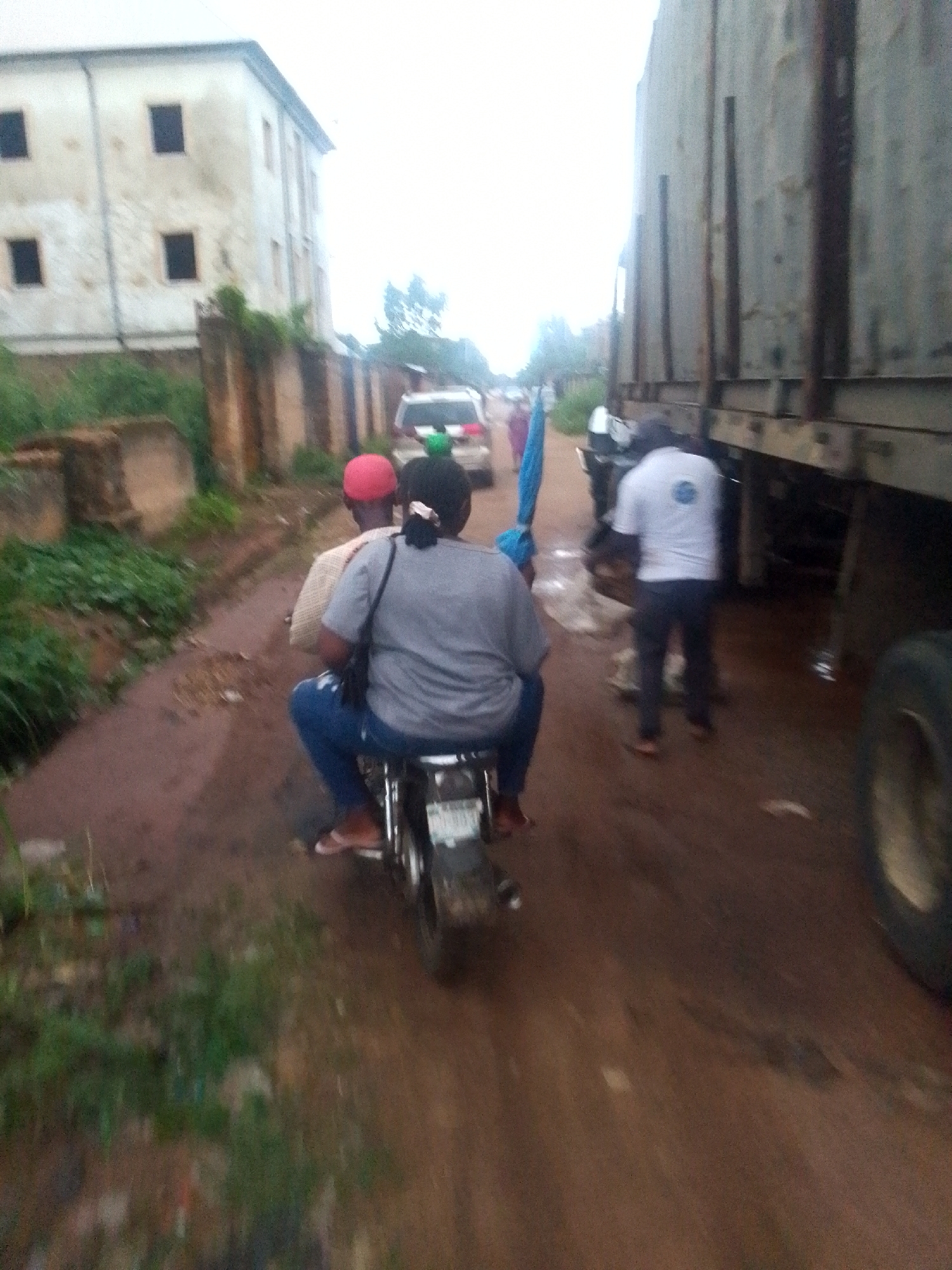 A blurry, low-resolution photograph depicts a dirt road in an unspecified location. A motorcycle with two passengers is moving away from the camera. The passenger in the back is wearing a gray shirt and blue jeans. The driver is wearing a red cap and light-colored shirt. To the right of the motorcycle is a large truck. A man in a white shirt and dark pants stands near the truck. In the background, other individuals and vehicles are visible. On the left, there is a dilapidated building and overgrown vegetation.