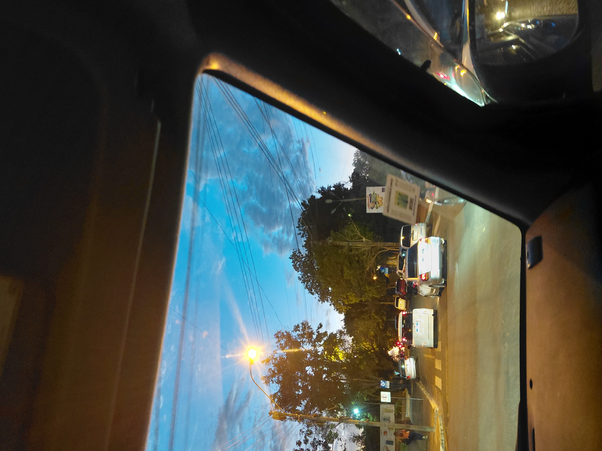 The image captures a view from inside a vehicle in Encarnación, Paraguay, looking forward through the windshield. The time appears to be dusk or early evening, with a twilight sky in shades of blue and hints of clouds. A street light with a bright yellow glow illuminates the scene on the left side of the road. Trees line the street, their foliage dark against the sky.
The road ahead shows moderate traffic, with several cars visible, including a white minivan and a silver sedan in the foreground, stopped at what appears to be an intersection or a pedestrian crossing marked by white stripes. Some of the cars have their headlights on. On the left sidewalk, a person in an orange shirt is walking away from the camera. Road signs, including one with a blue "E" symbol, are present on the left. The interior of the car is visible in the foreground, with the dashboard and a portion of the side pillar. The image is shot from a low angle, looking out through the car's windows.