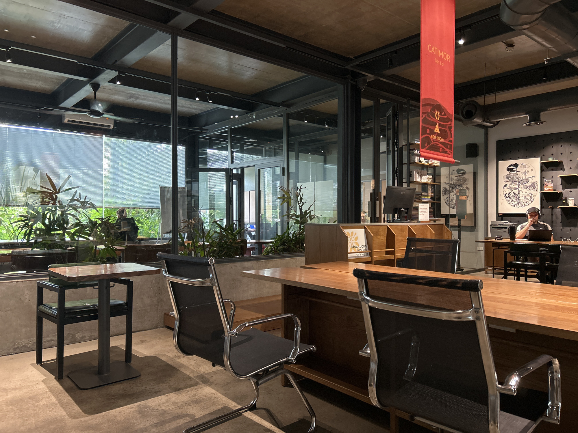 The image shows the interior of an office space in Da Nang, Vietnam. The office has a modern industrial design with exposed beams and concrete ceilings. Large windows with blinds look out onto lush greenery. In the foreground, there are several sleek, black mesh office chairs with chrome accents, positioned around polished wooden desks. One small table with a wooden top and black metal legs stands near the window. In the background, a man is seated at a desk, wearing headphones and working on a laptop. A red banner with text and graphics hangs from the ceiling. Shelving units filled with books and decorative items are visible along the walls. The overall atmosphere appears to be a contemporary and functional workspace.