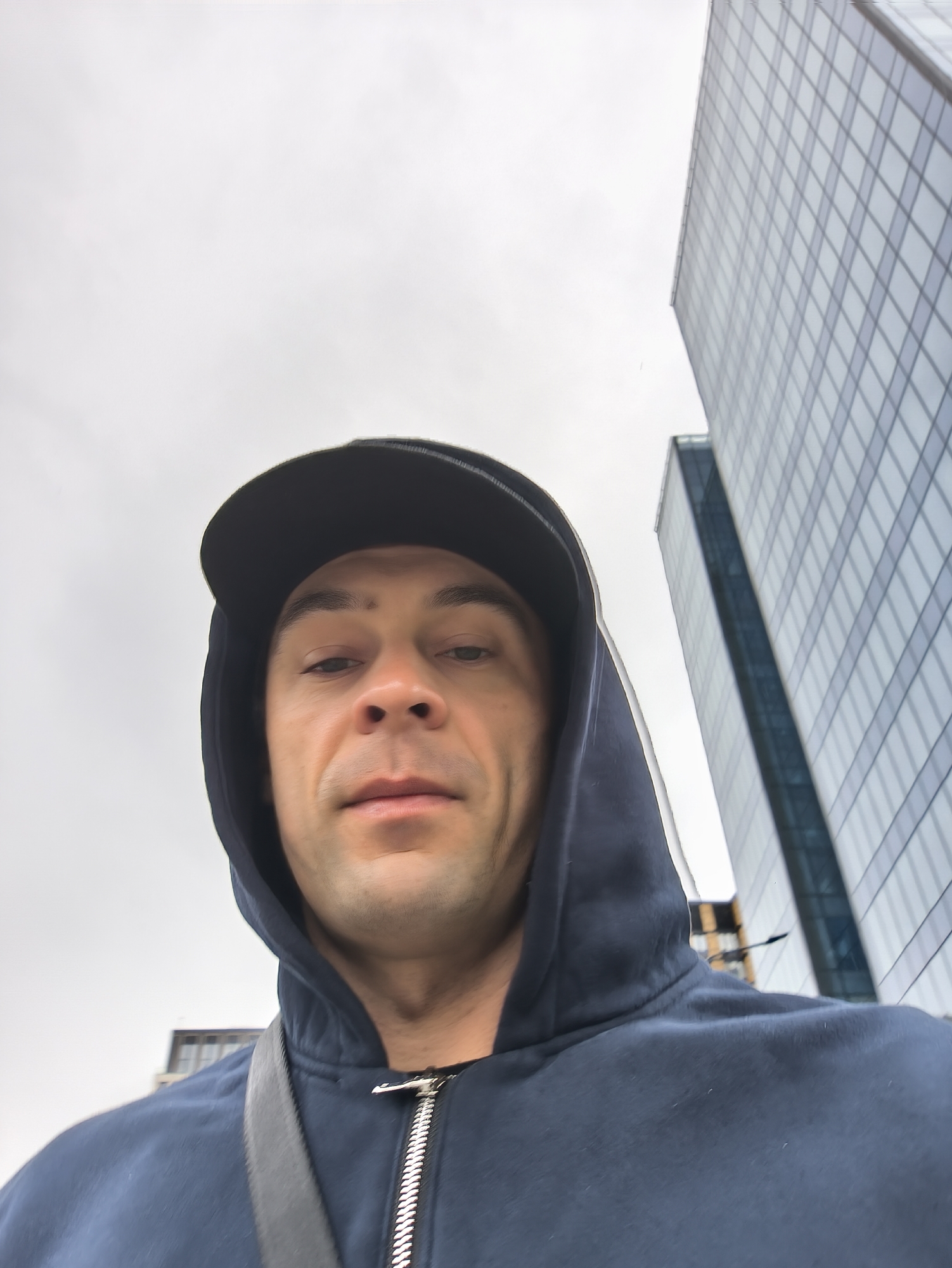 A man is taking a selfie outdoors. He is wearing a dark-colored hooded sweatshirt and a black baseball cap. A strap of some kind is visible over his shoulder. Behind him is a tall, modern glass skyscraper. The sky is overcast. The skyscraper suggests that the location is Moscow, Russia, specifically in the Moscow International Business Center known as Moscow-City.