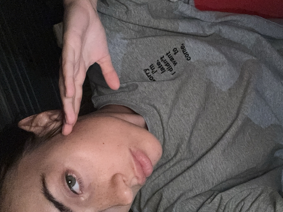 The image displays a close-up, angled portrait of a single individual, likely an adult female, positioned indoors. The subject has light skin, visible freckles on the nose and cheeks, light green eyes, and full lips. Dark hair is partially visible around the left ear. The individual is wearing a grey, short-sleeved t-shirt that features black text on the left chest area. The text reads: "sorry I'm late. I didn't want to come." The subject's left hand is raised, with fingers extended and pressed against the right side of the face, near the temple, partially obscuring the upper right facial area and ear. The gaze is directed towards the camera. The background is dark and out of focus, showing faint horizontal patterns behind the head, indicative of an indoor setting with subdued lighting. A small portion of red fabric or object is visible on the right periphery of the frame. The specific location within the city of Warsaw, Poland, cannot be determined from the visual information present in the image.