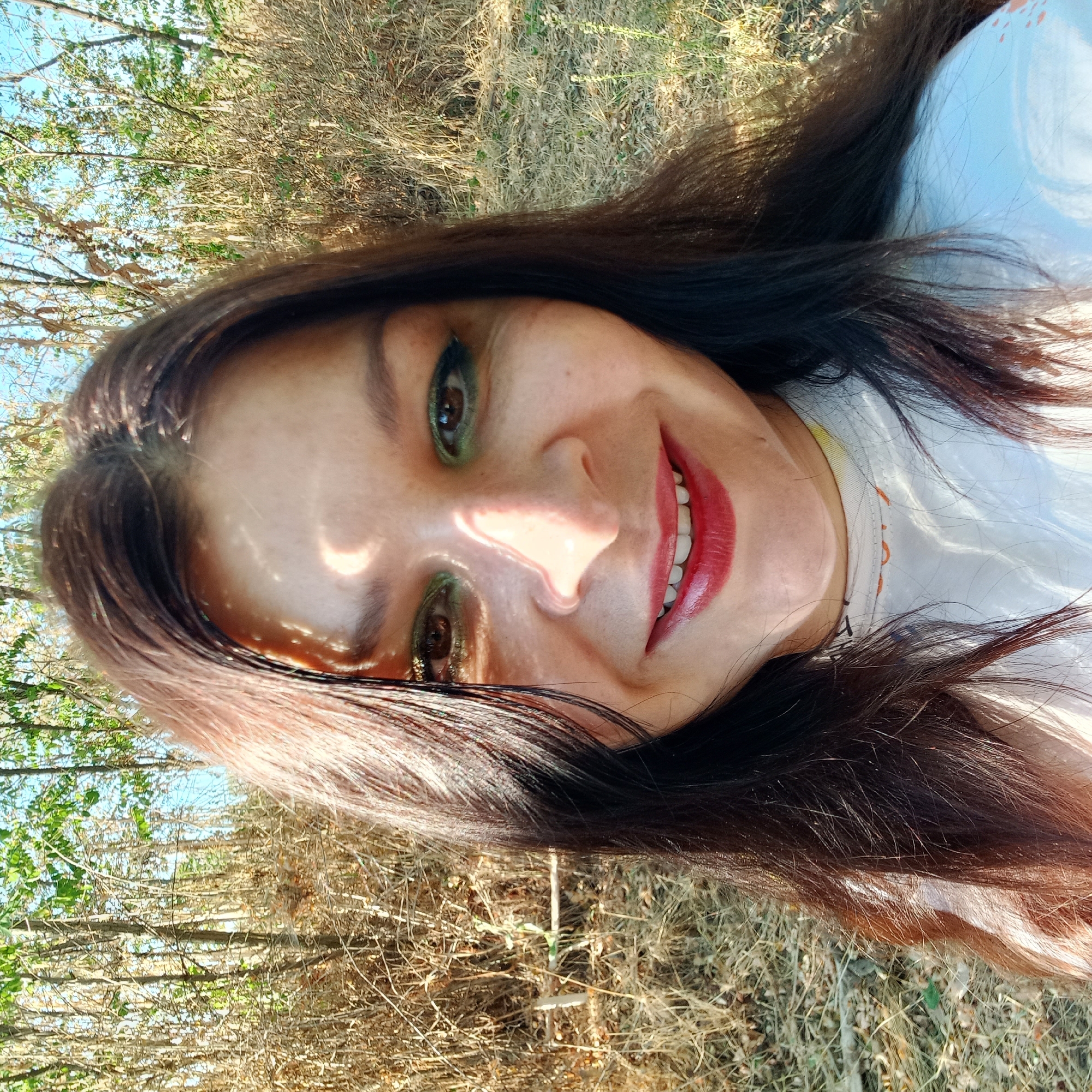 A woman with long, dark hair and green eyeshadow smiles at the camera, wearing a white t-shirt. She is standing outdoors in a dry, grassy area with trees in the background. Sunlight casts shadows on her face. The image does not provide any information that would allow for identification of a specific location in the city of Klyntsi, Ukraine.