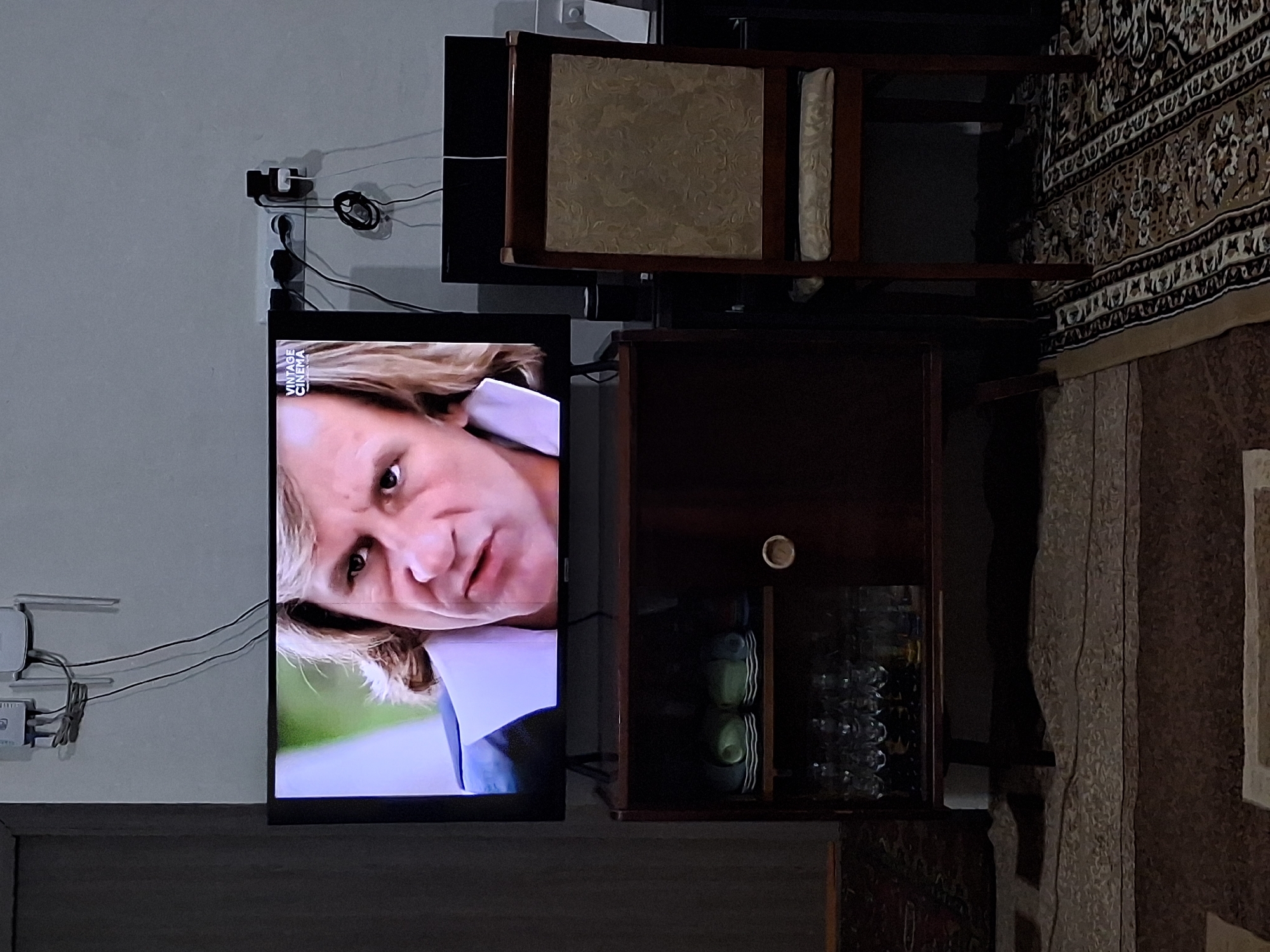 The image shows a living room scene. In the center, a flat-screen television displays a close-up of a man's face. The television is placed on top of a dark wooden cabinet. The cabinet has two open shelves, the top shelf containing stacked plates and cups, and the bottom shelf filled with glassware. To the right of the cabinet is a wooden chair with an upholstered seat and back. The room is sparsely decorated, with a dark rug covering the floor. Electrical outlets and wires are visible on the wall behind the television. The scene is set in Yeghegnadzor, Armenia.