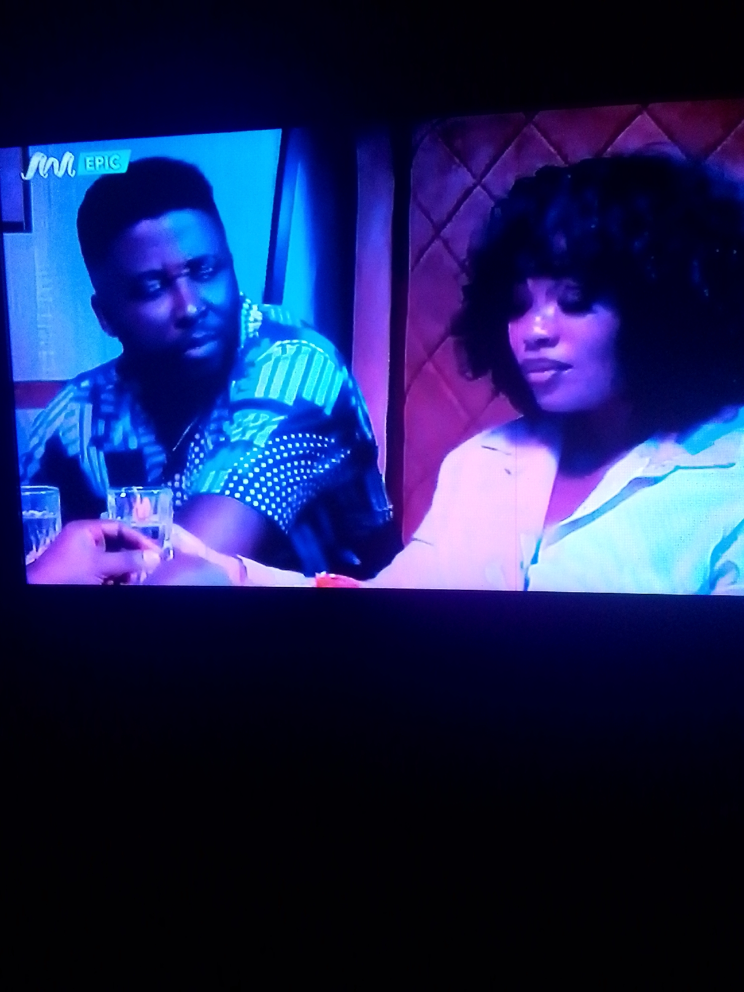 The media file shows two people sitting at a table, possibly in a dimly lit restaurant or lounge. A man with dark skin and short hair is on the left, wearing a patterned blue and white shirt. He is looking towards the right, with a neutral expression. In front of him are two glasses, one partially filled with a clear liquid. On the right is a woman with dark, curly hair and light skin, wearing a light-colored collared shirt. She is looking down and to her left, with a thoughtful or perhaps sad expression. Behind them, the wall is upholstered in a textured, quilted pattern, predominantly in shades of purple and pink, suggesting a sophisticated or intimate setting. The overall lighting is dim, with a blue and purple hue cast over the scene, creating a moody atmosphere. A small logo with the letters "M" and "EPIC" is visible in the upper left corner, indicating the content is from a media channel or production. The time of day is likely evening or night, given the lighting conditions.