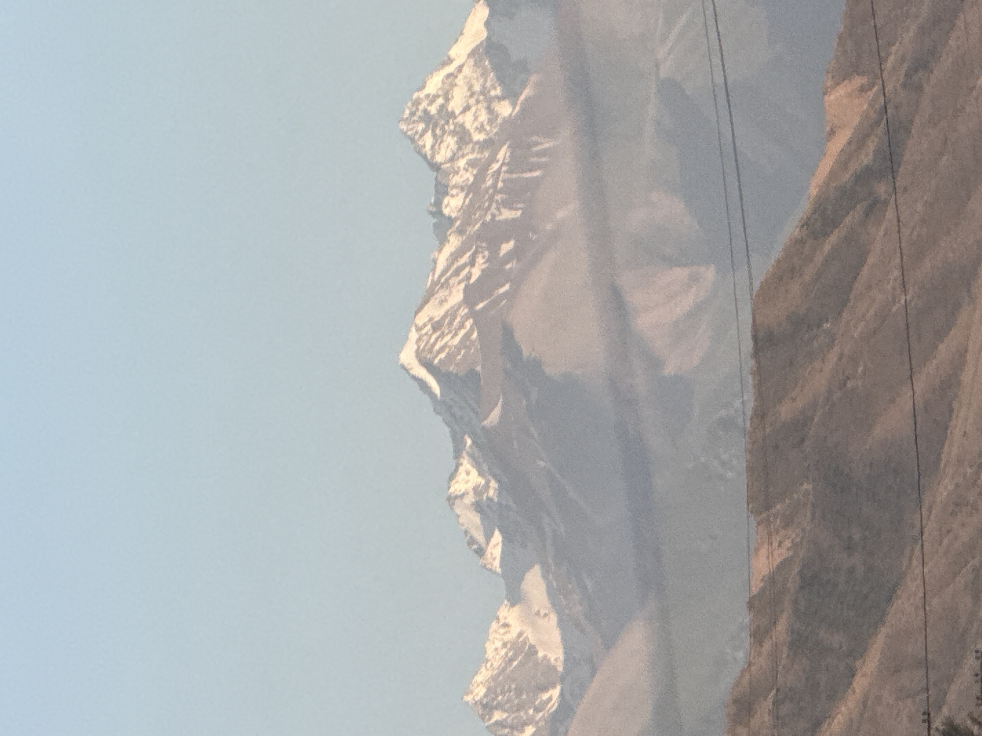 The image depicts a mountainous landscape under a clear, light blue sky. The mountains are rugged and appear to be snow-capped, with sharp peaks and dramatic slopes. The light suggests it is daytime, likely morning or afternoon, with the sun illuminating the snow-covered areas and casting shadows on the darker rock faces.
On the right side of the image, a steep, rocky incline is visible, sparsely vegetated with dry brush or grass. Several thin, dark lines, possibly power lines or cables, stretch across the frame, suggesting human infrastructure in the vicinity. These lines are partially obscuring the view of the mountains.
The overall impression is one of a vast, natural environment, perhaps a remote or high-altitude region. The scale of the mountains and the clear sky evoke a sense of grandeur and isolation. There are no people or specific objects that are clearly identifiable beyond the natural elements and the power lines. The image appears to be a photograph taken from a distance, possibly from a vehicle or a viewpoint overlooking the terrain. There is no visible text in the image.