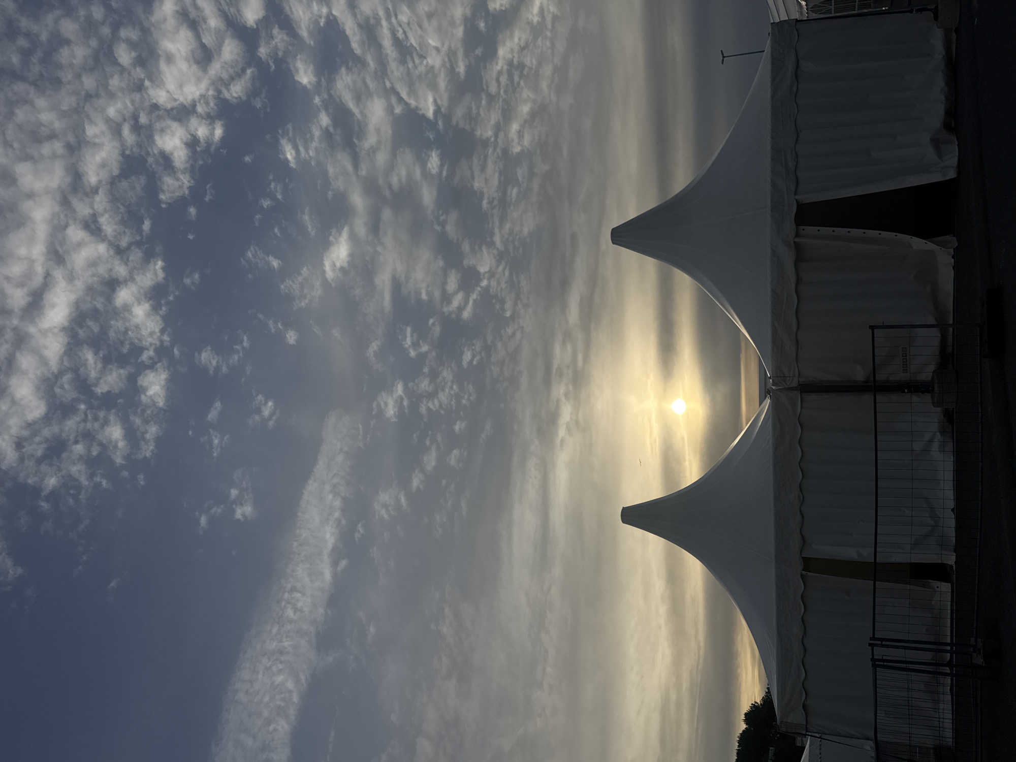 The image depicts a scene at dusk in Tallinn, Estonia, with the sun low on the horizon and casting a soft glow. In the foreground, two large, white, conical-roofed tents stand side-by-side, with a fence in front of them. The tents are taut and appear to be made of fabric. One tent has an open entrance, revealing a dark interior. The sky above is filled with scattered clouds, some wispy and others more dense, in shades of gray and white, with the sun appearing as a bright orb just above the horizon line. The overall atmosphere is serene and somewhat dramatic due to the lighting conditions. The surrounding environment appears to be an outdoor venue or event space. There are no people visible in the image.