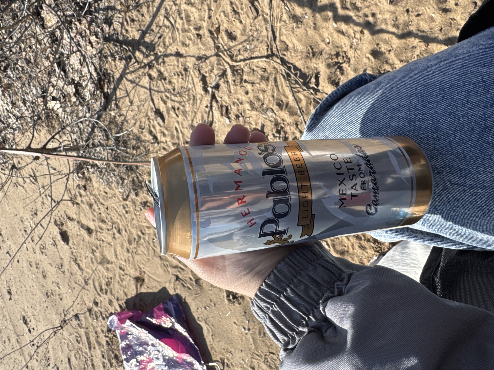 A person sits on a sandy surface, likely a beach or desert, holding a can of "Pablos Light Beer" with the label oriented horizontally. The person is wearing jeans and a grey jacket. The background shows dry twigs and sandy ground. A small, patterned bag lies on the sand near the can. The location is Unknown, Unknown.