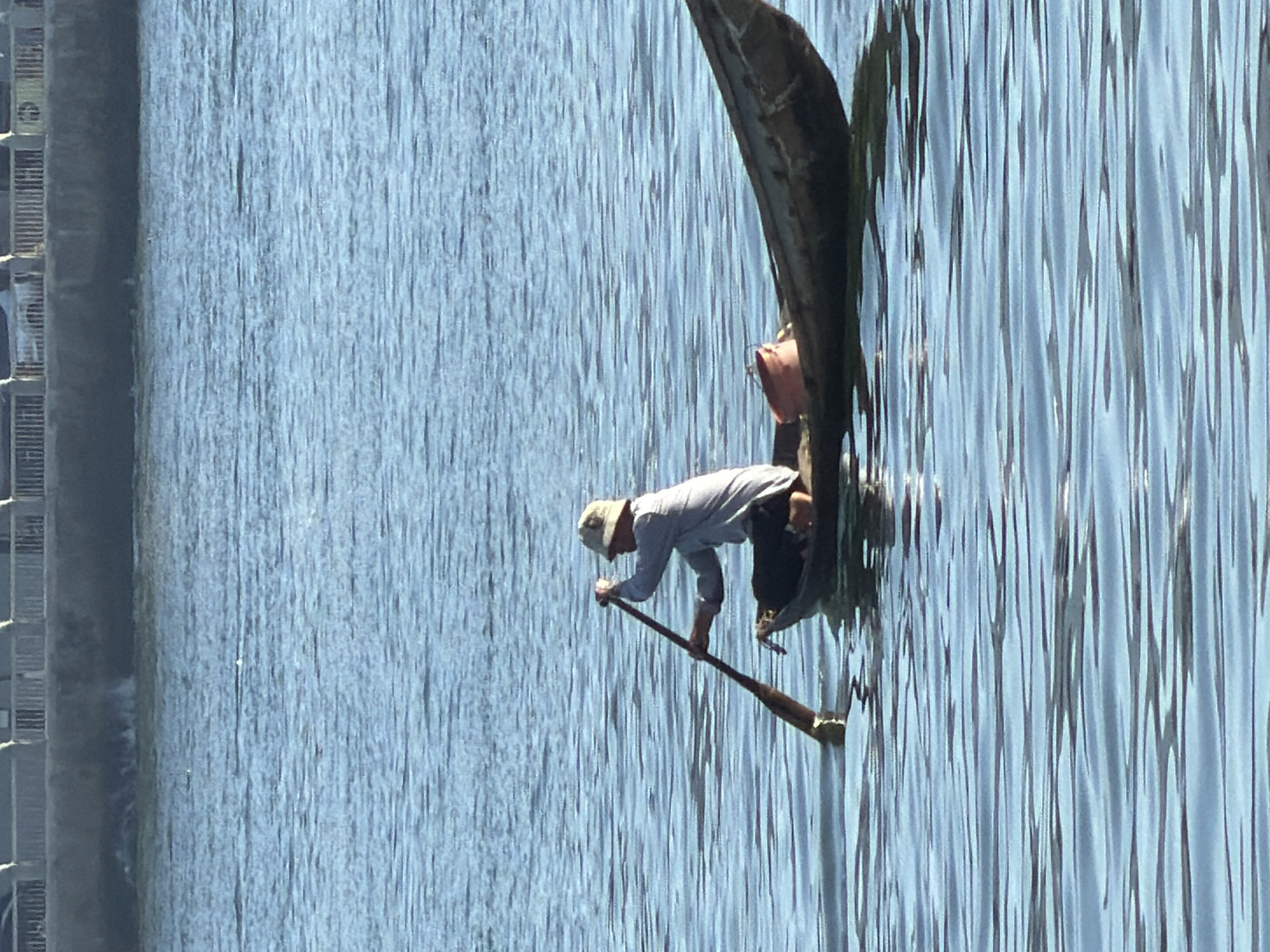 A person wearing a light-colored hat and shirt is rowing a small boat in the water. The boat is dark-colored and appears to be made of wood. There is a small orange bucket in the boat. The water is blue and appears to be calm with small ripples. In the background, there is a concrete structure with a railing, possibly a walkway or barrier along the waterfront, with some vehicles visible behind it. This scene is likely located in Nha Trang, Vietnam, given the characteristic appearance of the waterfront and the type of boat used for fishing or transportation.