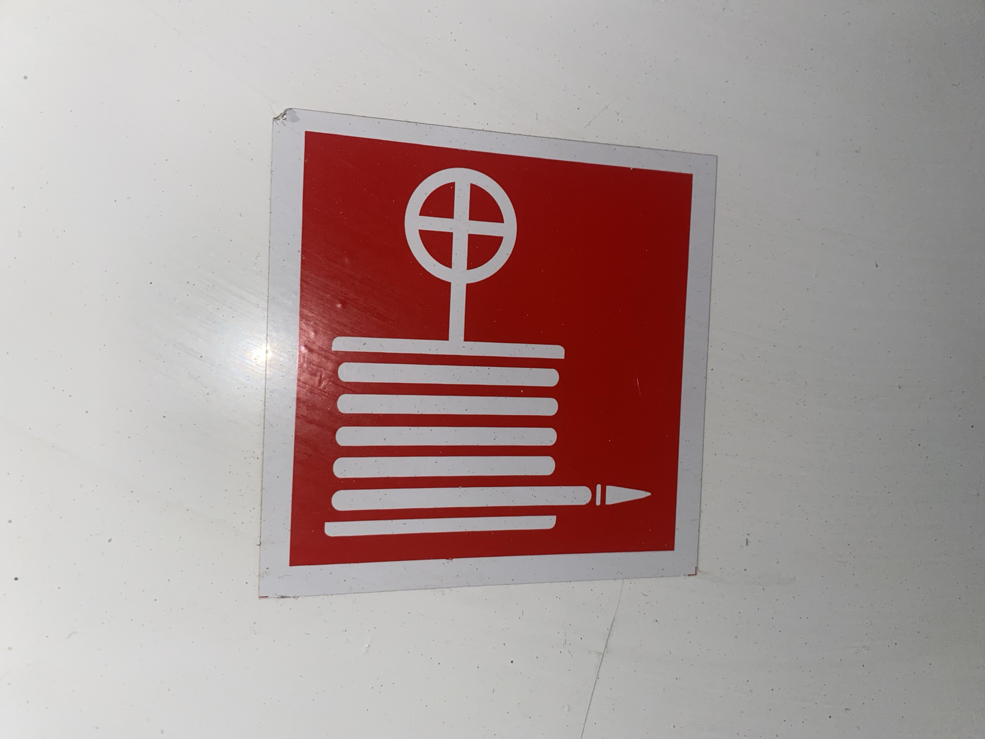The image shows a square red and white sign attached to a white surface. The sign depicts a white wheel-shaped symbol at the top, connected by a vertical line to a stack of six horizontal white lines. The bottom line extends to the right and terminates in a pointed shape, resembling a nozzle or spray pattern. The sign is slightly damaged at the top corner. The surface it's attached to appears to be slightly textured or speckled. Location and city information are not determinable from the image itself.