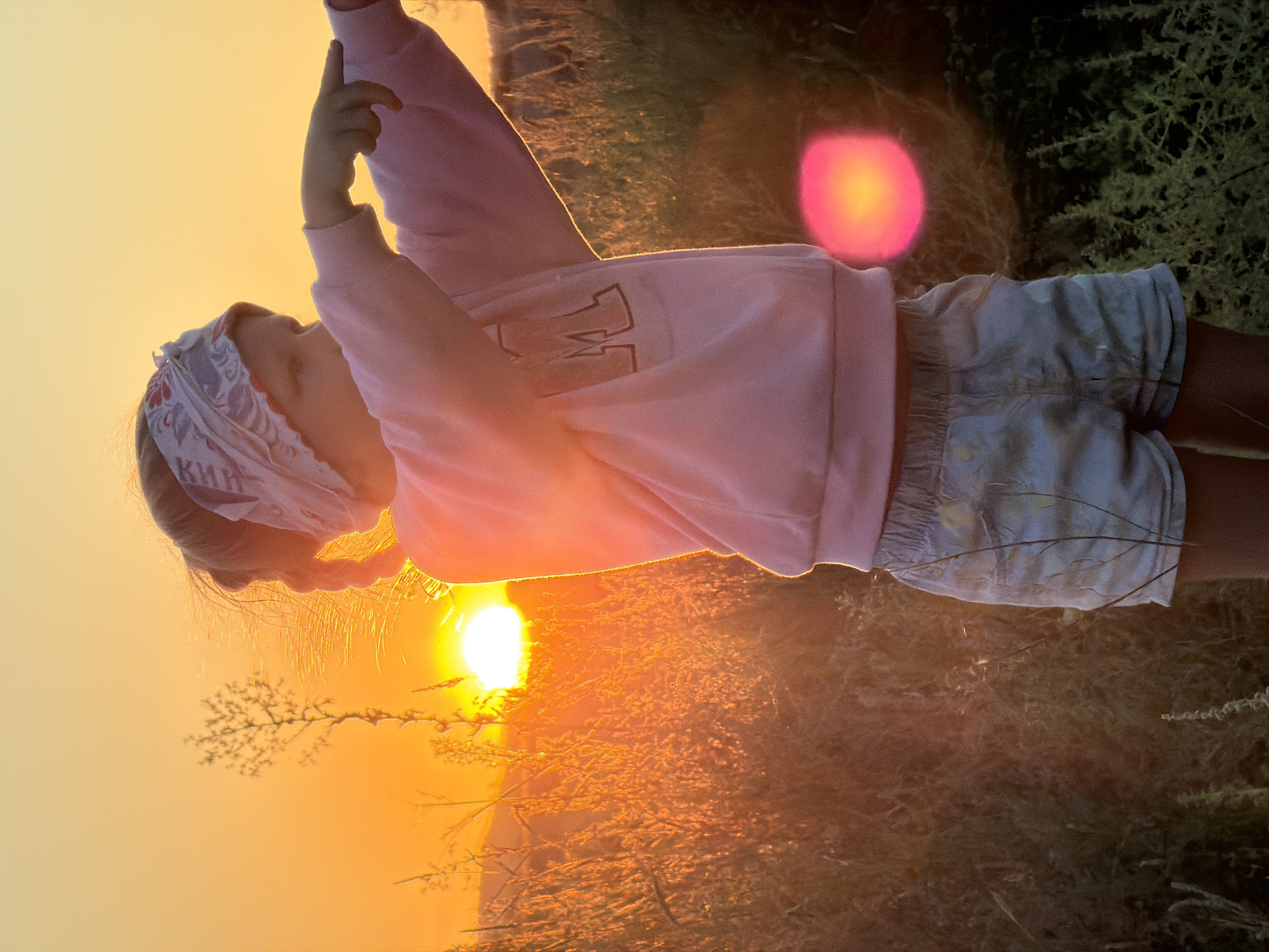 In Nizhnyaya Lipovka, Russia, a young girl stands in a field of dry grass at sunset. She is wearing a pink sweatshirt and light blue shorts, with a patterned bandana tied around her head. Her right arm is extended, and her index finger is pointing towards the setting sun, which is a bright orange orb low on the horizon. The light from the sun casts a warm glow over the scene, illuminating the tall grass and creating lens flare. The background is a hazy orange and yellow sky.