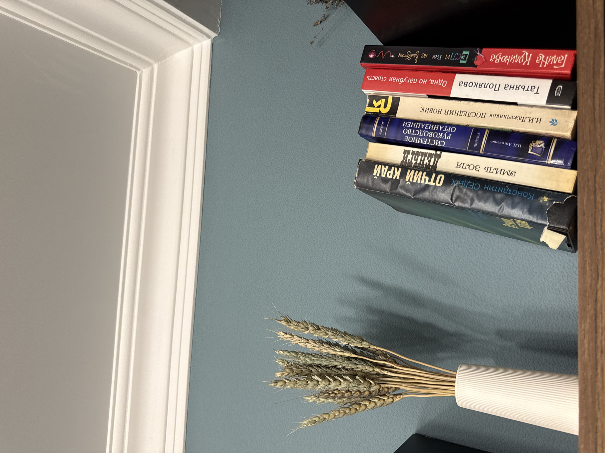 The image shows a stack of books on a shelf against a blue wall, with a white decorative trim to the left. The top book has a red cover with the title "Галина Куликова" and "Если вы не влюблены". The second book has a white cover with the title "Татьяна Полякова" and "Одна, но пагубная страсть". Another book titled "И.И.Лажечников ПОСЛЕДНИЙ НОВИК" and the one titled "СИСТЕМНОЕ РУКОВОДСТВО ОРГАНИЗАЦИЕЙ" are among others. The bottom book is dark with the title "Константин СЕДЫХ ОТЧИЙ КРАЙ". Below the books is a white vase containing a bundle of wheat stalks. No information about Ovoshchnoy, Russia can be gathered from the image.