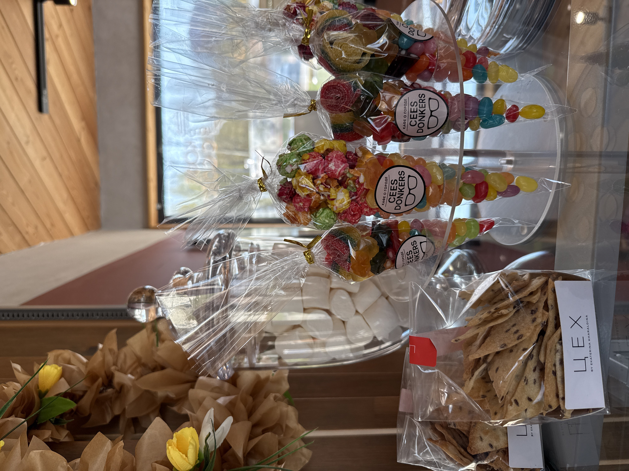 The image shows a display of packaged food items, seemingly inside a cafe or shop. There are four bags of mixed candies labeled "CEES DONKERS" with a logo that includes a pair of glasses and the text "КАФЕ О ГОРОДЕ", which translates to "Cafe in the City". Next to the candies are some bags of flaxseed crackers with a label that reads "ЦЕХ BY EKATERINA KHARALDINA". In the background, there are marshmallows in a glass container. Below the display, there's a decoration of brown paper and yellow and white artificial flowers. Based on the text "КАФЕ О ГОРОДЕ" and "CEES DONKERS", it is possible that the location is in the city of Votkinsk, Russia.
