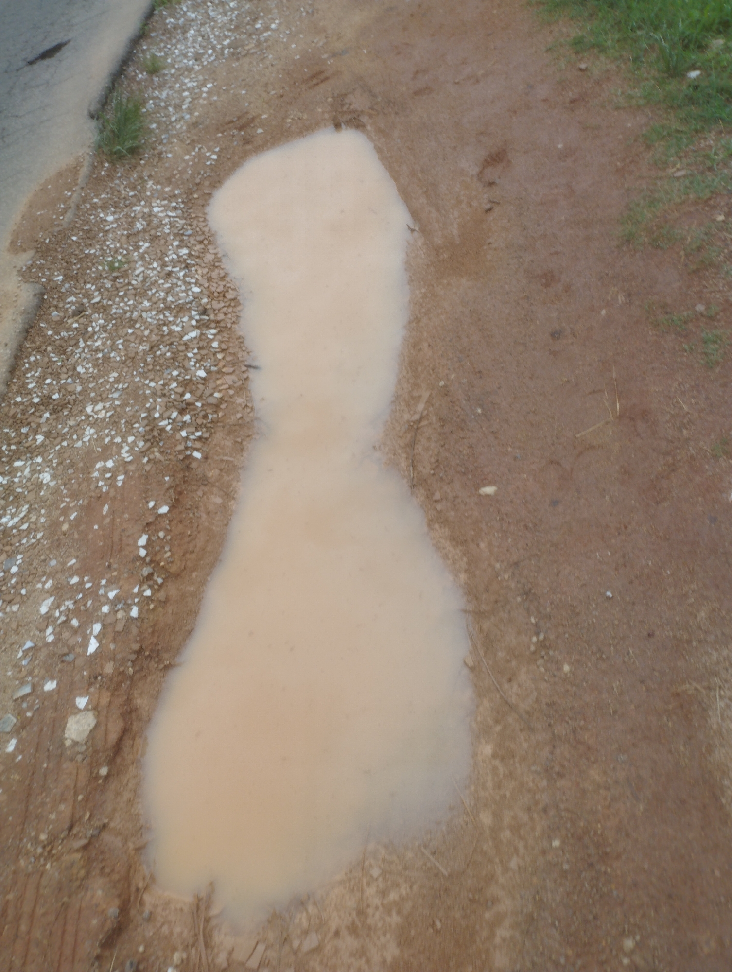 The image captures a muddy puddle on a dirt road, likely in Jos, Nigeria. The puddle, shaped somewhat like an hourglass, has a light brown, murky appearance, suggesting it's a mixture of water and soil. The road itself is composed of reddish-brown dirt and scattered gravel. To the left of the puddle, a section of a paved surface, possibly a road or sidewalk, is visible, with some lighter colored gravel and a patch of green grass. The overall environment appears to be an unpaved or partially paved area, common in many parts of Jos. No people or specific objects are clearly identifiable within the scene. The lighting suggests it's daytime, possibly overcast or late afternoon, given the diffused light and muted colors.