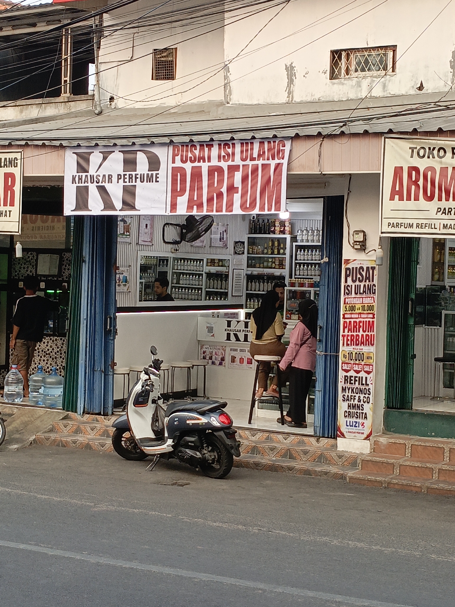 A street-level, eye-level, medium shot captures the storefront of a perfume shop located in East Jakarta, Indonesia. The shop, named "KHAUSAR PERFUME", displays banners advertising its products. One banner, in red and white, reads "PUSAT ISI ULANG PARFUM" (Refill Perfume Center), with "KHAUSAR PERFUME" written in large, stylized letters above it. Another banner, to the right, has "TOKO PARFUM AROMA" (Aroma Perfume Shop) in red lettering.
Inside the shop, shelves are lined with small bottles, presumably filled with perfumes. Two women are seated on stools at a counter, and one woman is standing. A male employee is visible behind the counter. In front of the shop, a man stands near several large water jugs. A white and blue scooter is parked on the street. The shop entrance is covered by blue and green corrugated metal shutters. The street in front of the shop is paved, and some of the stairs leading to the shop are visible. The buildings above the shop are two stories, with windows and visible electrical wiring. The sky appears to be a light gray, suggesting it may be late afternoon or early evening.