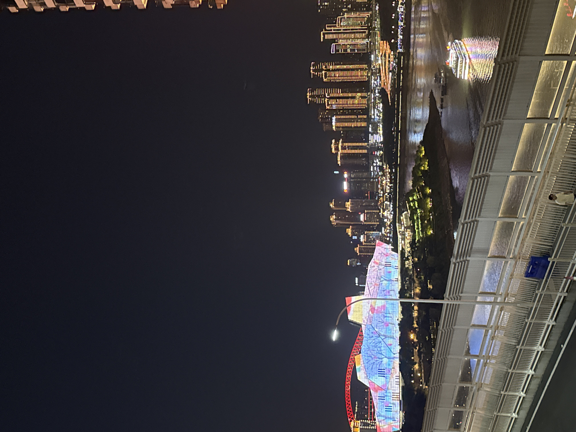 This is a nighttime photograph taken in Yuzhong District, China, featuring a cityscape and a river. The image is oriented vertically, with the top of the frame showing the lower portion of a building and the bottom showing a bridge.
The scene depicts a vibrant urban environment at night. On the right side of the frame, a modern bridge with white railings and illuminated pathways is prominent. The bridge appears to be elevated, offering a view of the river and the city skyline beyond. A few people are visible on the bridge.
Across the river, a dense city skyline is illuminated with countless lights. Tall buildings with glowing windows stretch across the horizon. On the river itself, a boat is brightly lit, with its colorful reflections shimmering on the water's surface.
In the foreground on the left, a large, uniquely designed building or structure is lit up with colorful patterns, predominantly in shades of blue and pink, with some red accents. A street lamp is also visible in front of this illuminated structure.
The overall impression is one of a lively and modern city at night, with the river serving as a central element connecting the different parts of the scene. The time of day is clearly night due to the darkness of the sky and the extensive artificial lighting. The weather appears clear, as evidenced by the sharp details of the city lights and their reflections.