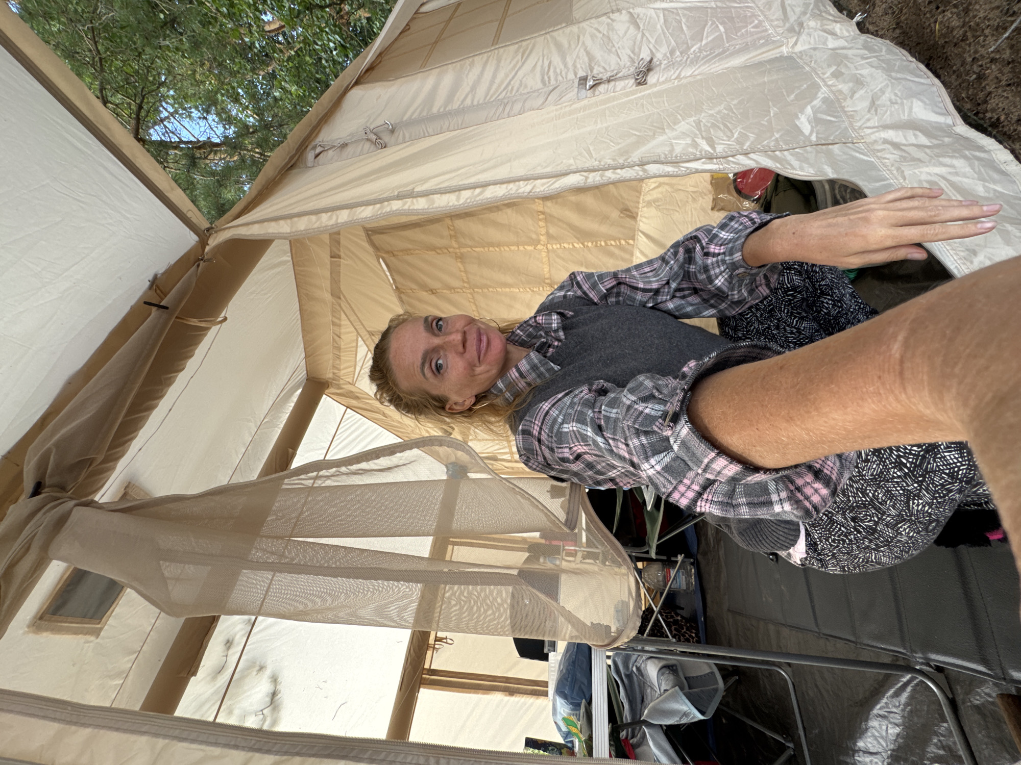A candid, eye-level, full-body shot shows a Caucasian woman with blonde hair pulled back, fair skin, and light freckles posing inside a large, beige canvas tent. She is wearing a grey v-neck sweater vest over a pink and grey plaid long-sleeved shirt, and patterned black and white pants. Her right arm is extended towards the camera, holding the phone for the selfie. The tent's interior is visible, with mesh windows and fabric partitions. Outside the tent, trees and blue sky suggest an outdoor environment, possibly a campsite in Zelenaya Roshcha, Russia, given the context of the query. Several camping chairs and tables are visible around the tent.