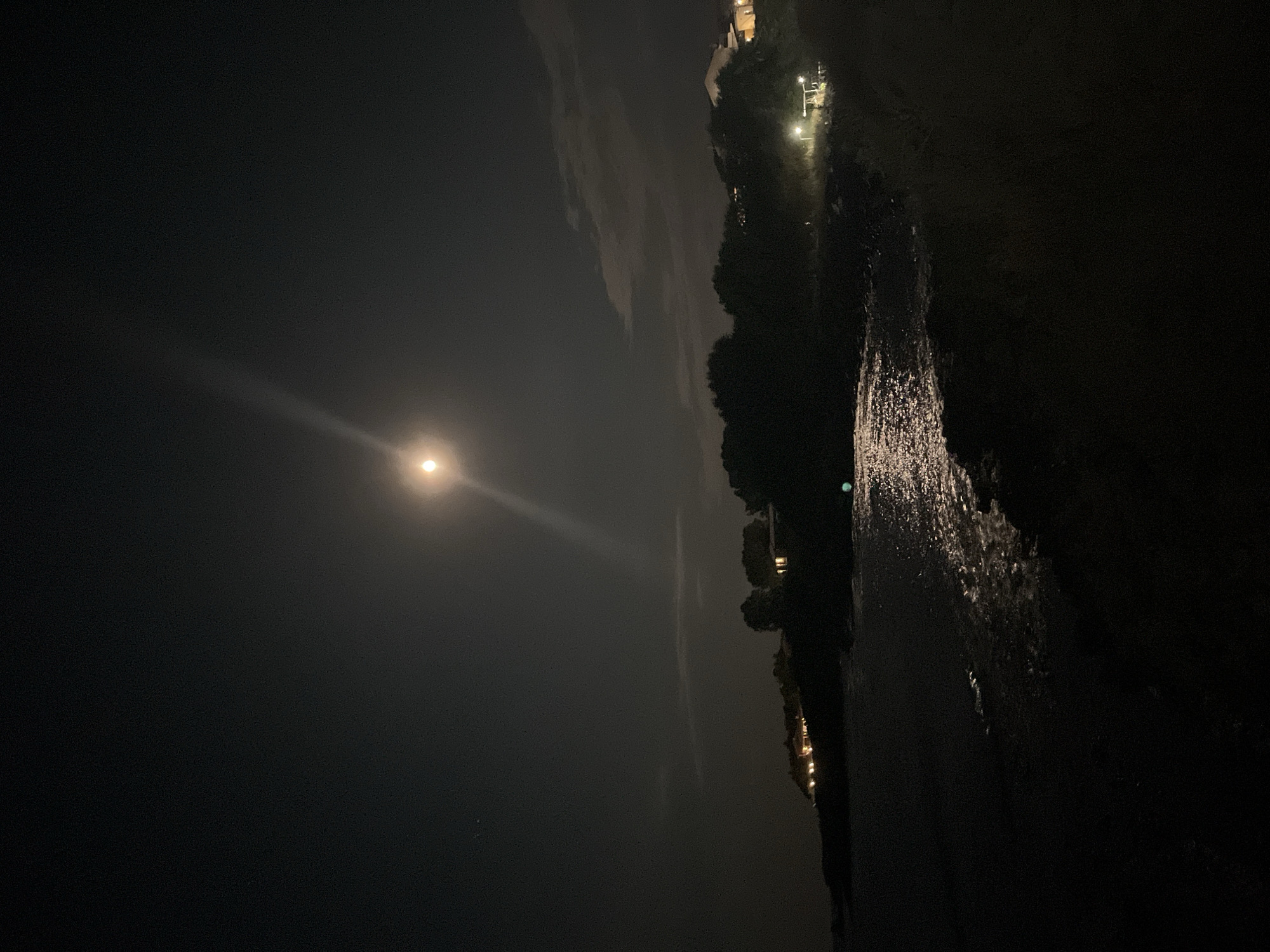 This is a nighttime photograph of the coast in Lozenets, Bulgaria. The moon is visible in the upper center of the frame, casting a bright reflection on the water. The sea is dark with small waves lapping at the shore. On the left side of the image, houses with illuminated windows are nestled on a hill overlooking the water. Trees are silhouetted against the night sky. The scene is dark and peaceful, with the moon and the lights from the houses providing the primary illumination.