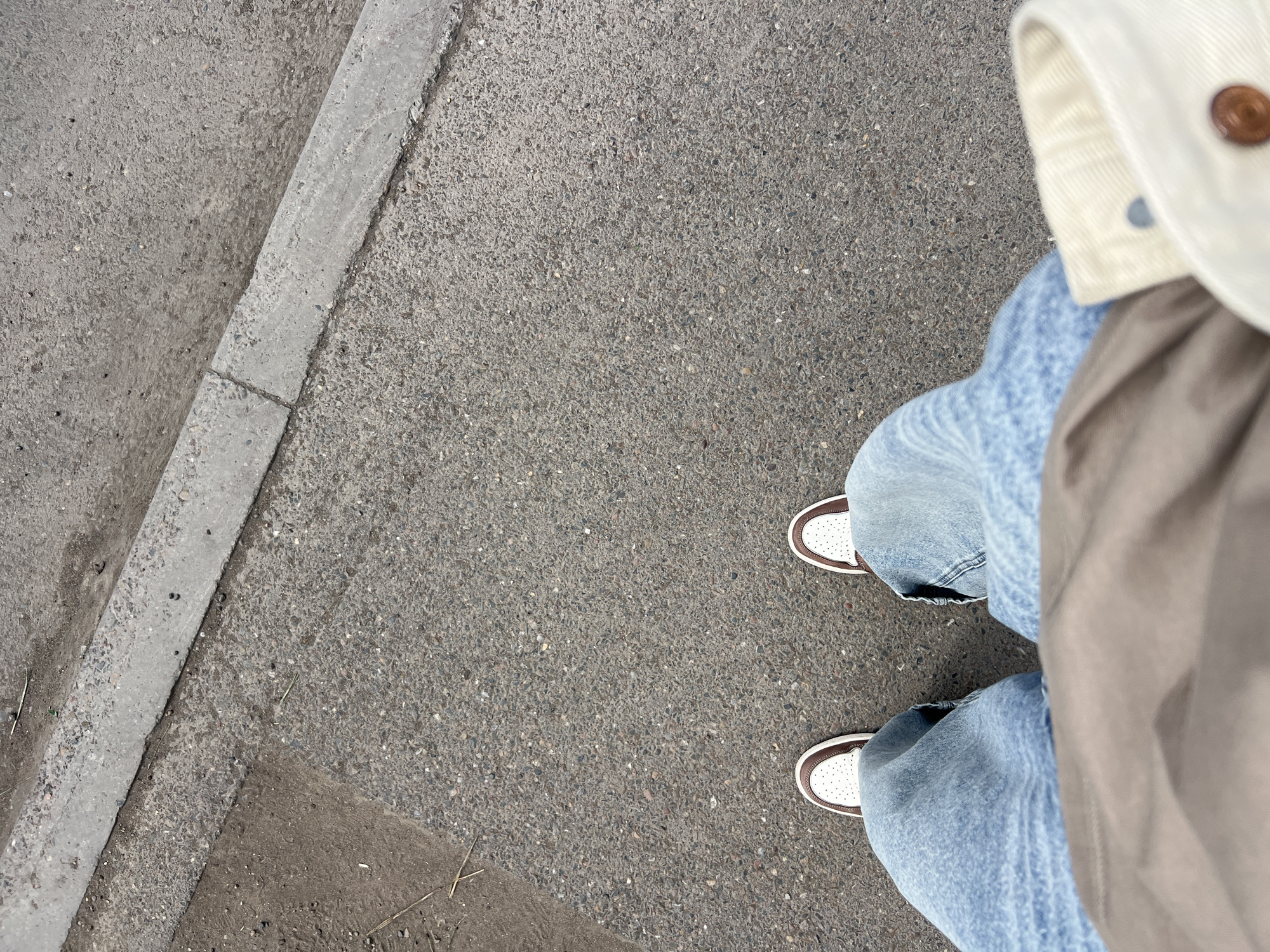 A high-angle shot shows a person standing on an asphalt surface in an urban environment. The person is wearing light blue jeans and white sneakers with brown trim. A portion of a white and brown jacket is also visible. To the left of the person is a concrete curb. The image was taken in Krasnoyarsk, Russia.