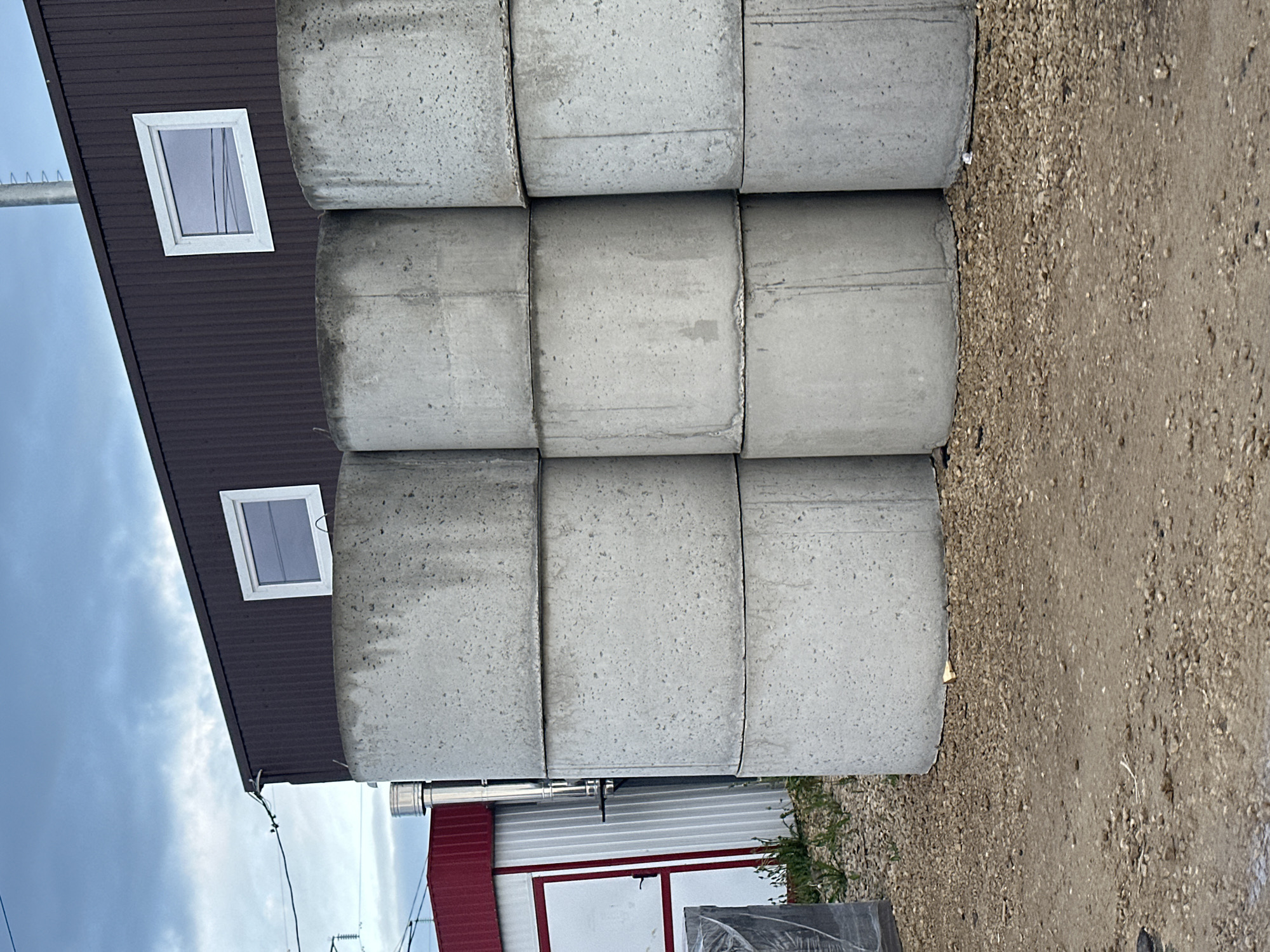 The scene depicts a stack of nine concrete rings, possibly for well construction, positioned next to a building in what appears to be a gravel or dirt lot. The building is a two-story structure. The upper portion is brown with two square windows. The lower portion is white with a red roofline. In the background, power lines are visible against a cloudy sky. The location is Эстачи, Russia.