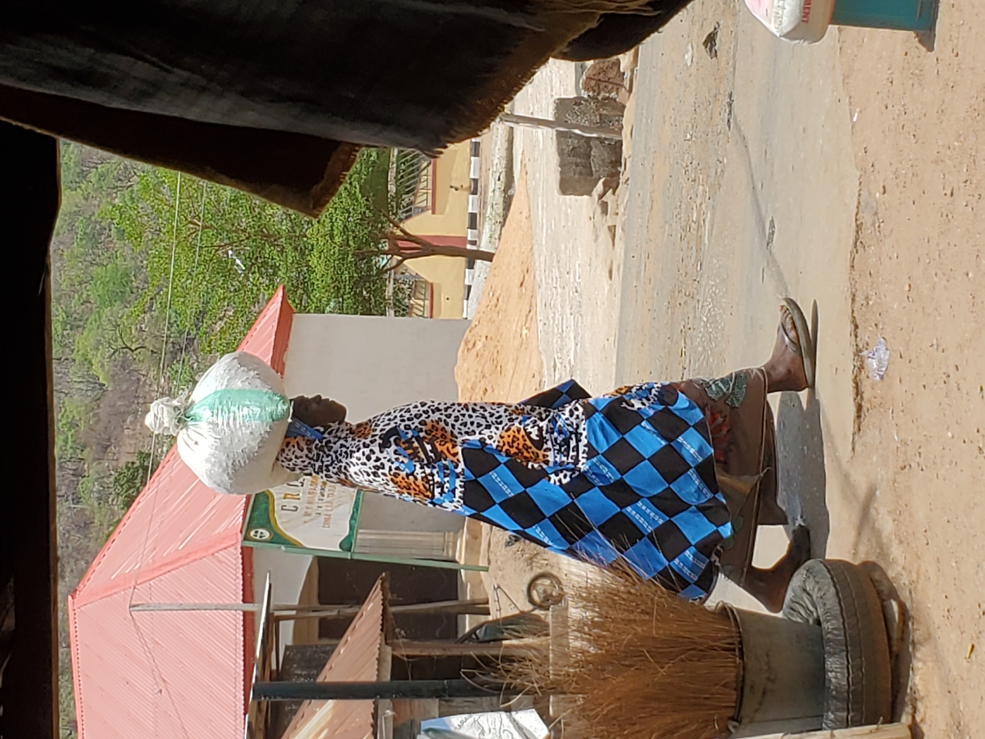 A woman carries a white sack on her head while walking on a paved road in Mararaba, Nigeria. She wears a dress with a blue and black checkered pattern on the bottom and a white, black, and orange spotted pattern on top. She is barefoot. The road is cracked and has patches of bare ground. A broom is visible in the foreground, next to a tire mounted on a metal stand. A building with a red roof and a sign is on the left. Trees and a hillside are in the background.