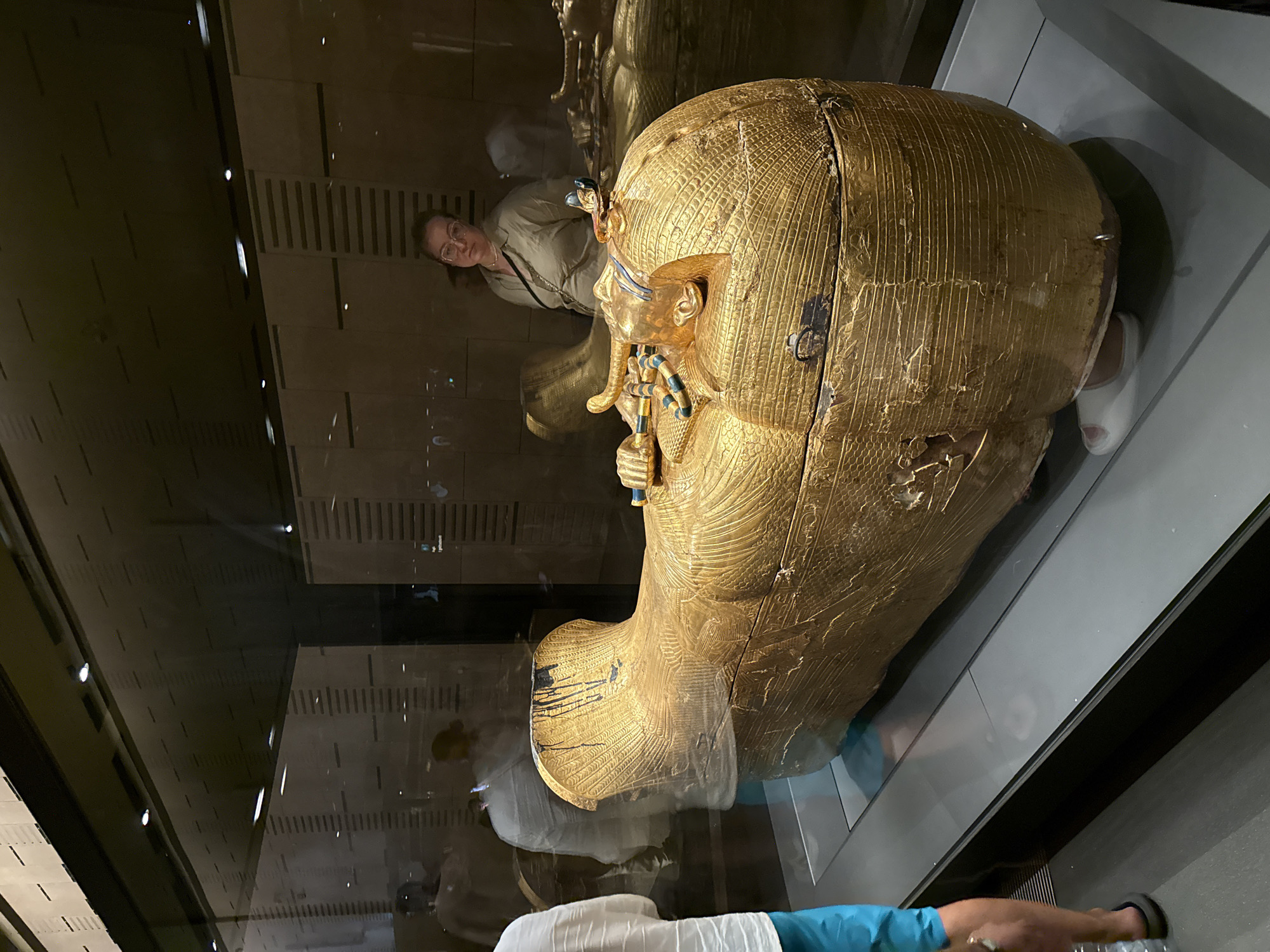In Kafrat Nassar, Egypt, a woman wearing glasses and a beige top stands behind a large, golden sarcophagus displayed in a museum. The sarcophagus is intricately decorated with patterns and appears to be made of gold. The lighting in the museum is dim, with spotlights directed at the sarcophagus. Reflections of people and the museum environment are visible on the glass case surrounding the artifact. A portion of another person's legs, wearing blue shorts and flip-flops, is visible in the foreground on the left side of the image. The background shows walls with horizontal paneling and further displays, suggesting a museum setting.