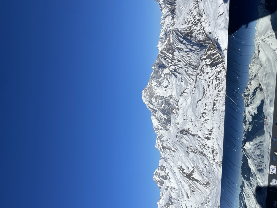 The media file presents a striking aerial or high-altitude view of a vast, snow-covered mountain range under a brilliant blue sky, likely in the French Alps near Séez. The image, if rotated 90 degrees counter-clockwise for correct orientation, shows the clear, intense blue sky dominating the upper half, with a subtle gradient from a deeper blue at the top to a slightly lighter hue towards the horizon.
The lower half of the image is filled with rugged, majestic mountains, heavily blanketed in pristine white snow. Exposed dark rock faces and dramatic peaks and ridges are visible, indicating deep valleys and a complex topography. The strong sunlight illuminates the snow, creating sharp contrasts and highlights that emphasize the textures and contours of the landscape.
In the right foreground, a dark, reflective surface, resembling a window or part of a vehicle interior (possibly an airplane or helicopter), is visible. This surface displays reflections of additional snow-covered terrain, extending the perceived depth of the mountainous scene. Within this reflective area, a small rectangular shape with faint white text "HP" is discernible, suggesting a laptop screen or a device label.
The scene depicts a bright, clear daytime with excellent visibility and sunny weather. There are no clouds in the sky, highlighting the vastness and serenity of the alpine environment. No people are visible, emphasizing the grandeur and untouched nature of the landscape. The overall impression is one of awe-inspiring natural beauty and immense scale.