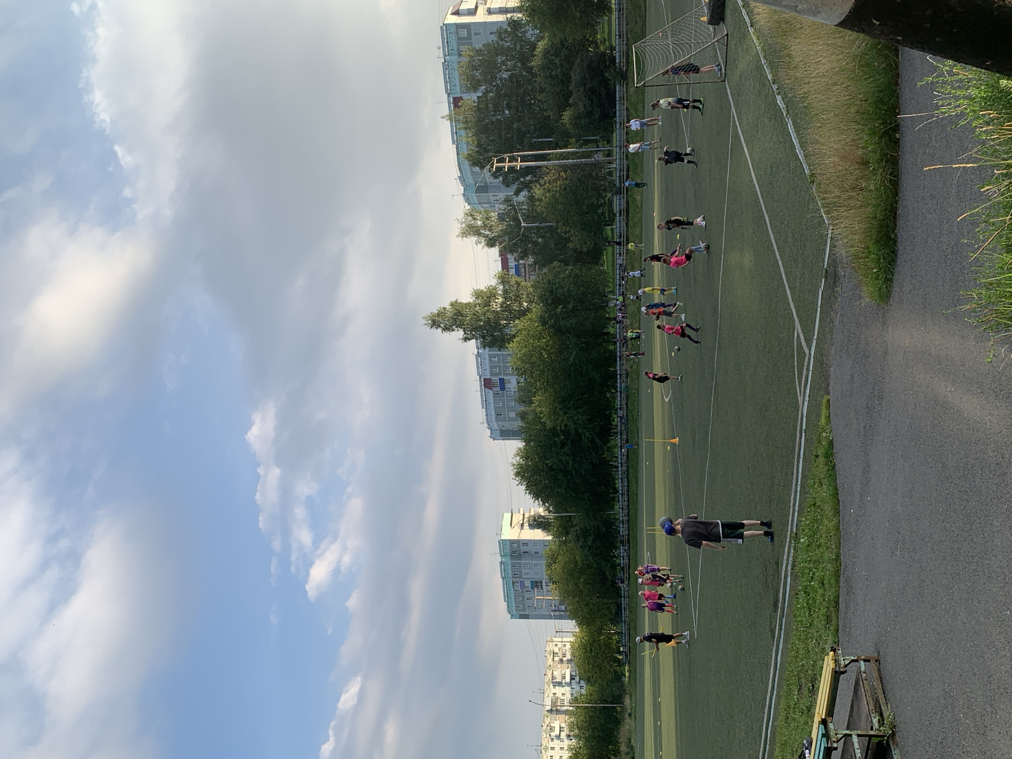 The image shows a wide shot of a green astroturf soccer field with several groups of children and some adults playing or observing. The field is marked with white lines and scattered with yellow cones. In the background, there are several tall, multi-story apartment buildings and a line of trees. The sky is partly cloudy with a warm, late afternoon light.
On the field, one group of children in pink shirts and dark shorts is gathered around a soccer ball, possibly receiving instruction. To their left, a boy in a dark shirt and shorts stands with two yellow sticks. Further to the right, another group of children is spread out, some with a soccer ball. A boy in a dark t-shirt and shorts, wearing a blue baseball cap, stands in the foreground looking towards the field. A soccer goal with a net is visible on the far right side of the field, with a person in a dark dress standing near it.
The scene appears to be a youth soccer practice or game in a residential area. The buildings in the background suggest an urban or suburban environment. The presence of apartment buildings and a sports field is characteristic of many residential districts in cities across Russia, including Prokopyevsk.