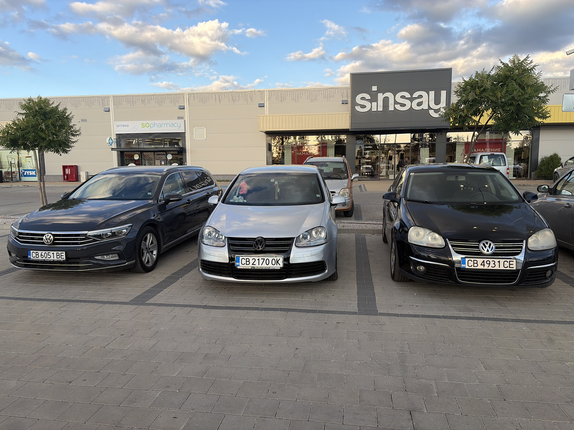 In Elin Pelin, Bulgaria, a parking lot is depicted in front of a commercial building under a partly cloudy sky. Three Volkswagen cars are parked: a dark gray Passat on the left, a silver Golf in the middle, and a black Golf on the right. The Passat has a license plate reading "CB 6051 BE." The silver Golf's license plate is "CB 2170 OK," and the black Golf's license plate is "CB 4931 CE." Behind the cars, the building features signage for "sinsay" and "SOpharmacy." Trees are present to the left and right of the building entrance. The parking lot is paved with interlocking gray bricks.
