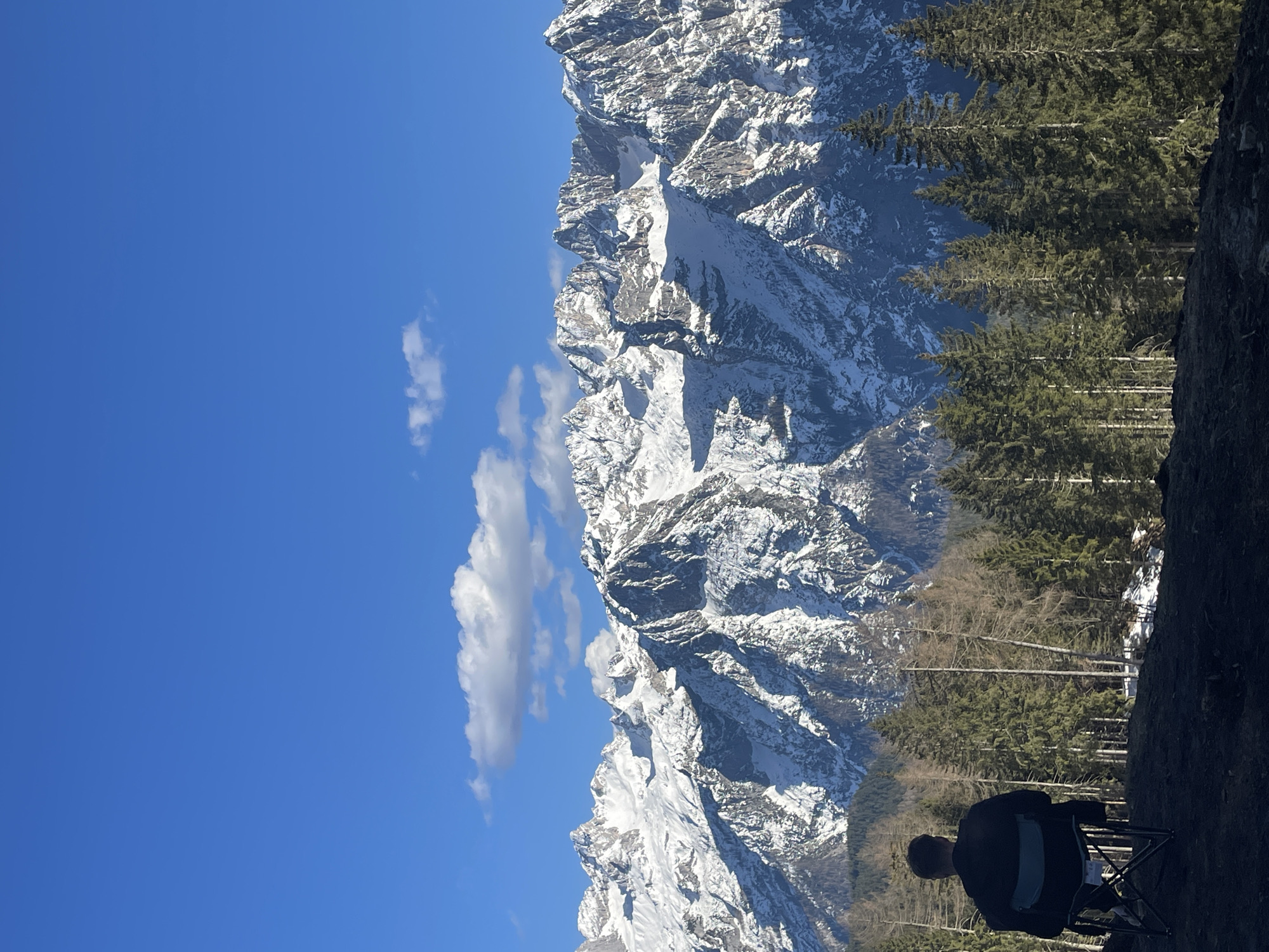 The image shows a mountain landscape in Cortina d'Ampezzo, Italy, with a clear blue sky and some white clouds. A large, snow-covered mountain range dominates the view. In the foreground, there are evergreen trees and a dark area, possibly a rock face. A person is sitting in a folding chair. The person is facing away from the camera.