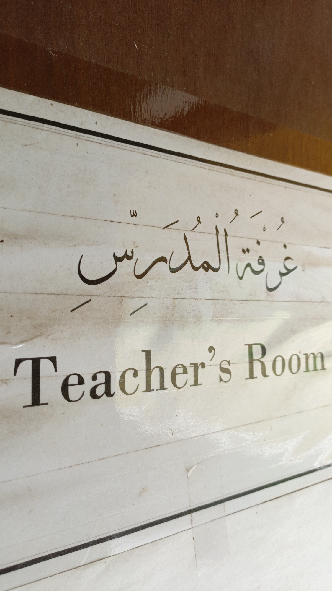 The image shows a sign with text in Arabic script and English. The Arabic text, which appears to be "غرفة المدرس", translates to "Teacher's Room". Below it, the English text "Teacher's Room" is clearly visible. The sign is mounted on a white surface with faint horizontal lines. The top portion of the image shows a brown, textured surface, possibly wood. The image is taken at an angle, with the sign slightly tilted. The scene does not provide any specific information about its location in the city of Trihanggo, Indonesia, or any details about people, actions, or interactions. It is a close-up of a sign.