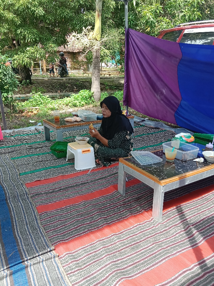 A woman wearing a hijab sits cross-legged on a colorful striped mat in an outdoor setting. She's holding food, possibly eating. Two small tables flank her, one displaying food containers and a beverage, the other with food. A small white stool is nearby. A purple and blue sheet hangs in the background, obscuring a vehicle. Trees and greenery are visible in the distance, along with a glimpse of people and a scooter. The scene appears to be a casual outdoor meal or gathering.