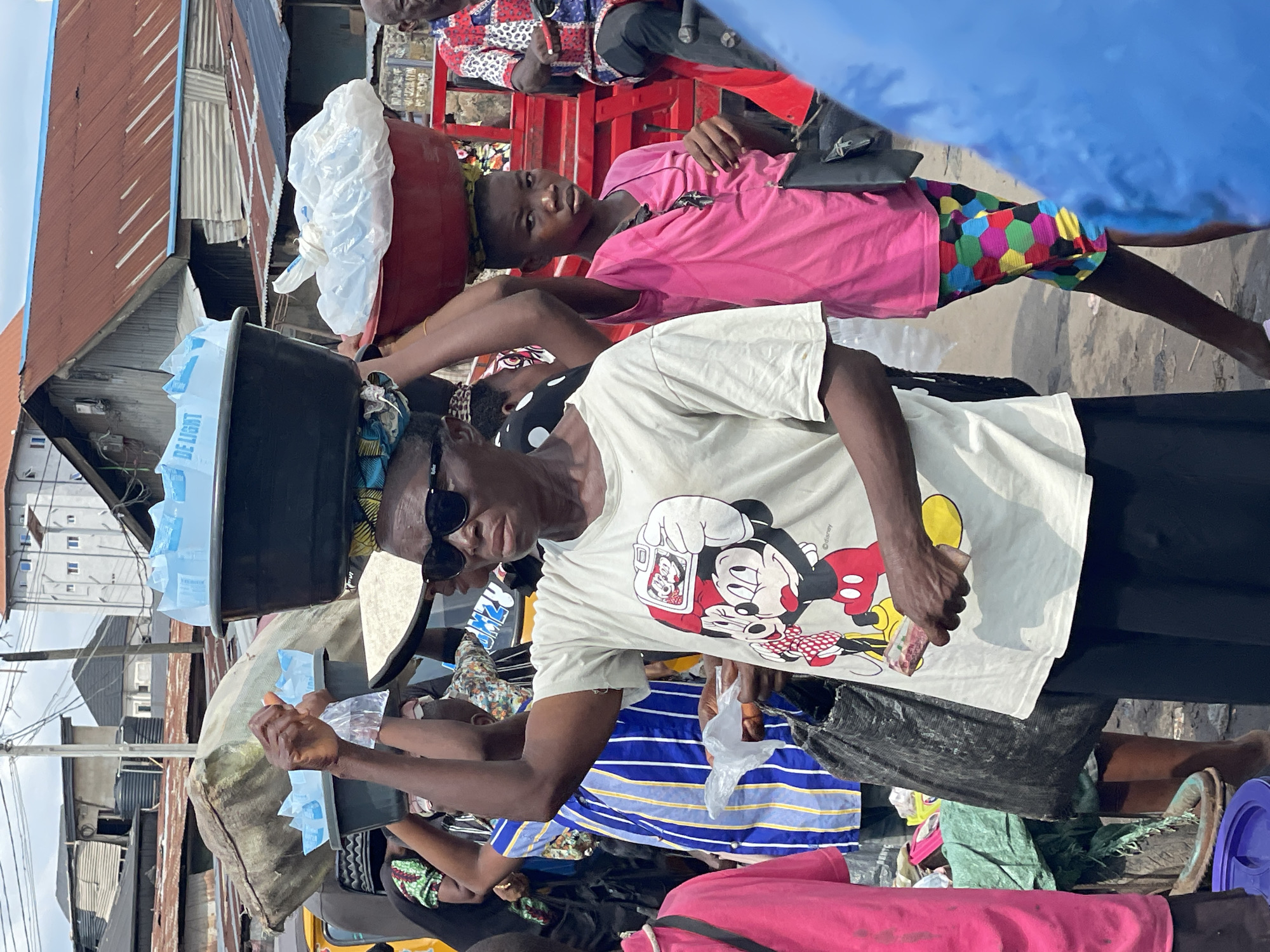 An outdoor, daytime scene captures a bustling street or market environment. In the foreground, a dark-skinned woman wears a light-colored t-shirt featuring a large Mickey Mouse graphic, black sunglasses, and dark pants. She balances a black plastic basin containing multiple light-blue packaged items, some labeled "DE LIGHT," on her head. Her right hand holds a stack of banknotes, and a black plastic bag hangs from her left shoulder. To her right, a young dark-skinned girl wears a bright pink t-shirt and multi-colored hexagonal-patterned shorts. She carries a red basin on her head, containing a crumpled white plastic bag, and a black shoulder bag. She looks directly forward with a neutral expression.
Various other individuals are partially visible: to the far left, a person in a pink t-shirt; behind the central woman, a person in a blue and white striped shirt also carries an item on their head and holds a small clear plastic bag. To the far right, an older man with a bald head wears a red, white, and blue patterned shirt and is seated on a red structure or vehicle.
The background features densely packed buildings with a mix of corrugated metal roofs, some rusted red, others gray. Multiple power lines crisscross the sky. Water tanks are visible on rooftops in the distance. The ground appears unpaved and uneven. A yellow vehicle is partially visible behind the central figures. The sky is overcast. The scene depicts active commerce, with people carrying goods, characteristic of a market or busy street in Unknown, Unknown.