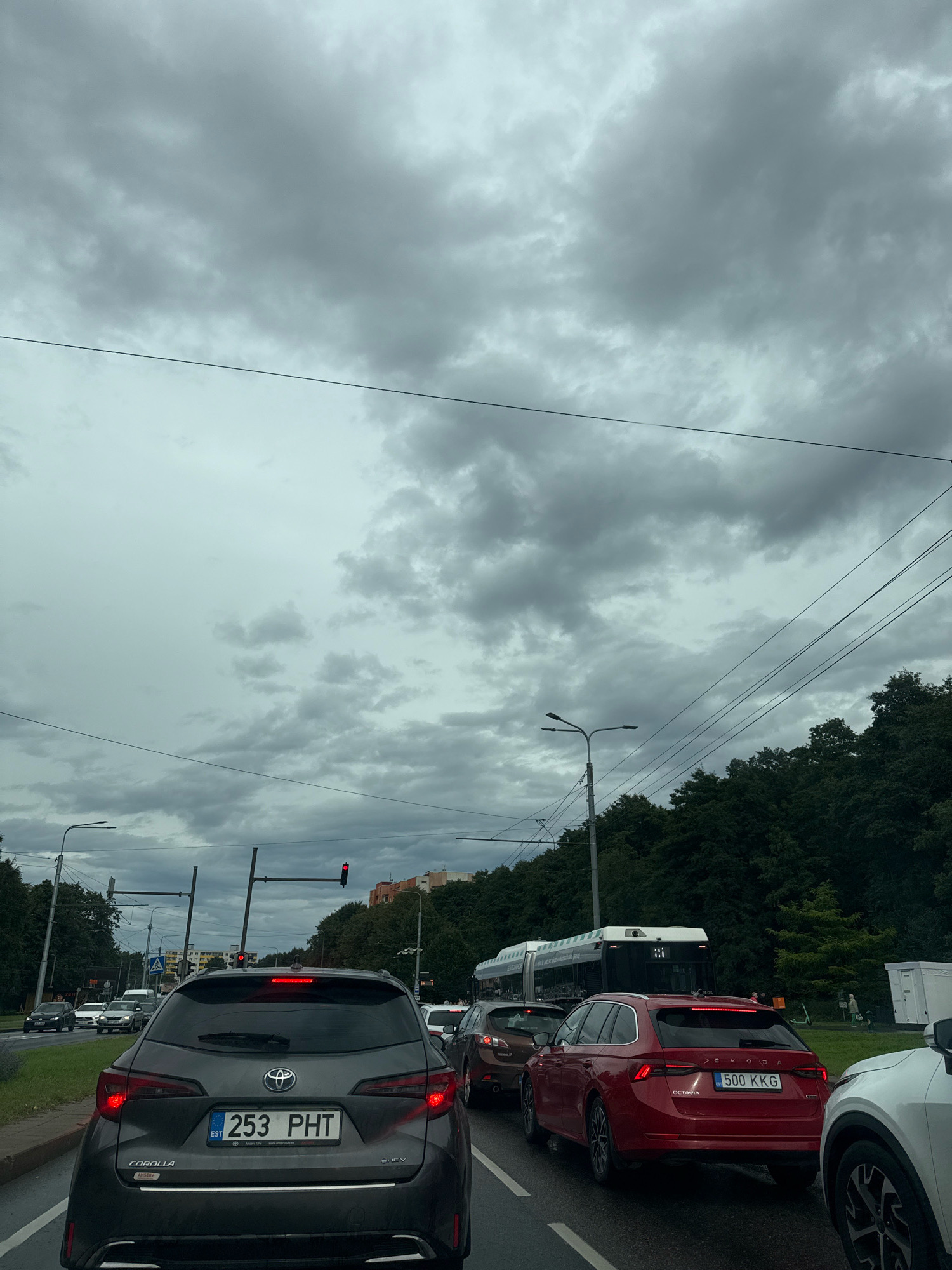 The image shows a traffic jam on a cloudy day in Tallinn, Estonia. In the foreground, a dark gray Toyota Corolla with license plate "253 PHT" is visible from the rear. To its right, a red Skoda Octavia with license plate "500 KKG" is also seen from the rear. Several other cars, including a white SUV on the far right, are also part of the traffic. A bus is seen in the middle ground. On the left side of the road, there are traffic lights, streetlights, and trees. In the background, buildings and more trees are visible. The sky is heavily overcast with gray clouds. The road appears wet, suggesting recent rain.