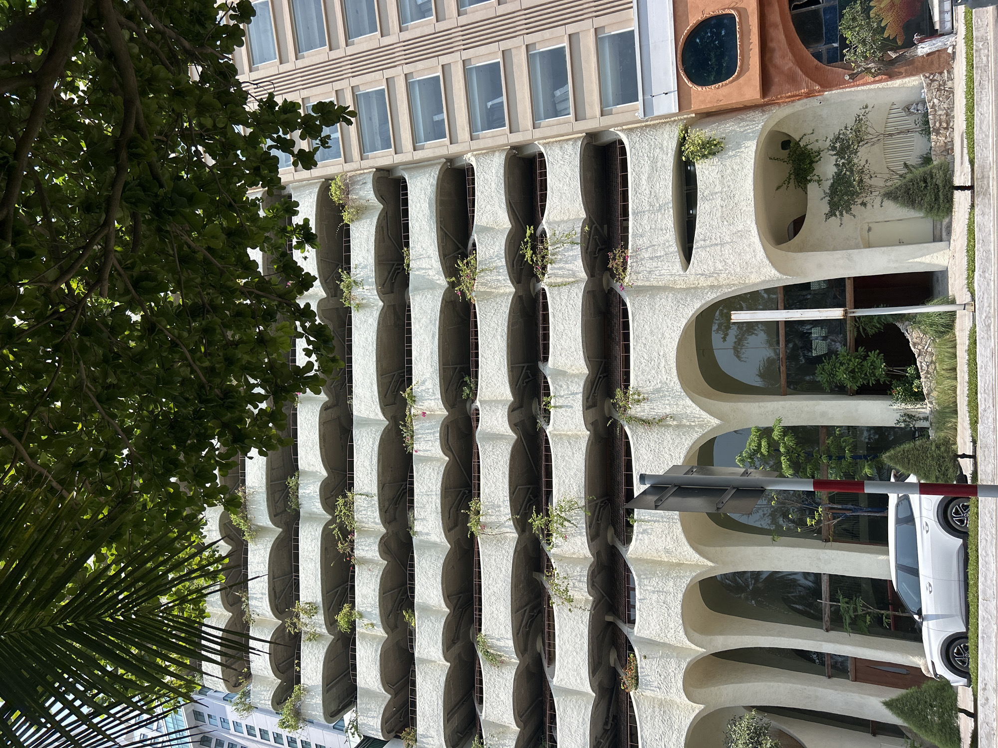 The photo captures a building facade in Bắc Nha Trang, Vietnam, exhibiting distinctive architectural features. The building is segmented horizontally with a clear distinction between the top and lower parts. The upper section features rectangular windows in a uniform pattern. Below, the facade transitions into a series of arched recesses with small plants sprouting from each opening. A tree with green leaves is present on the left, partially obscuring the view. A white car is parked near the base of the building. The light suggests it is daytime.