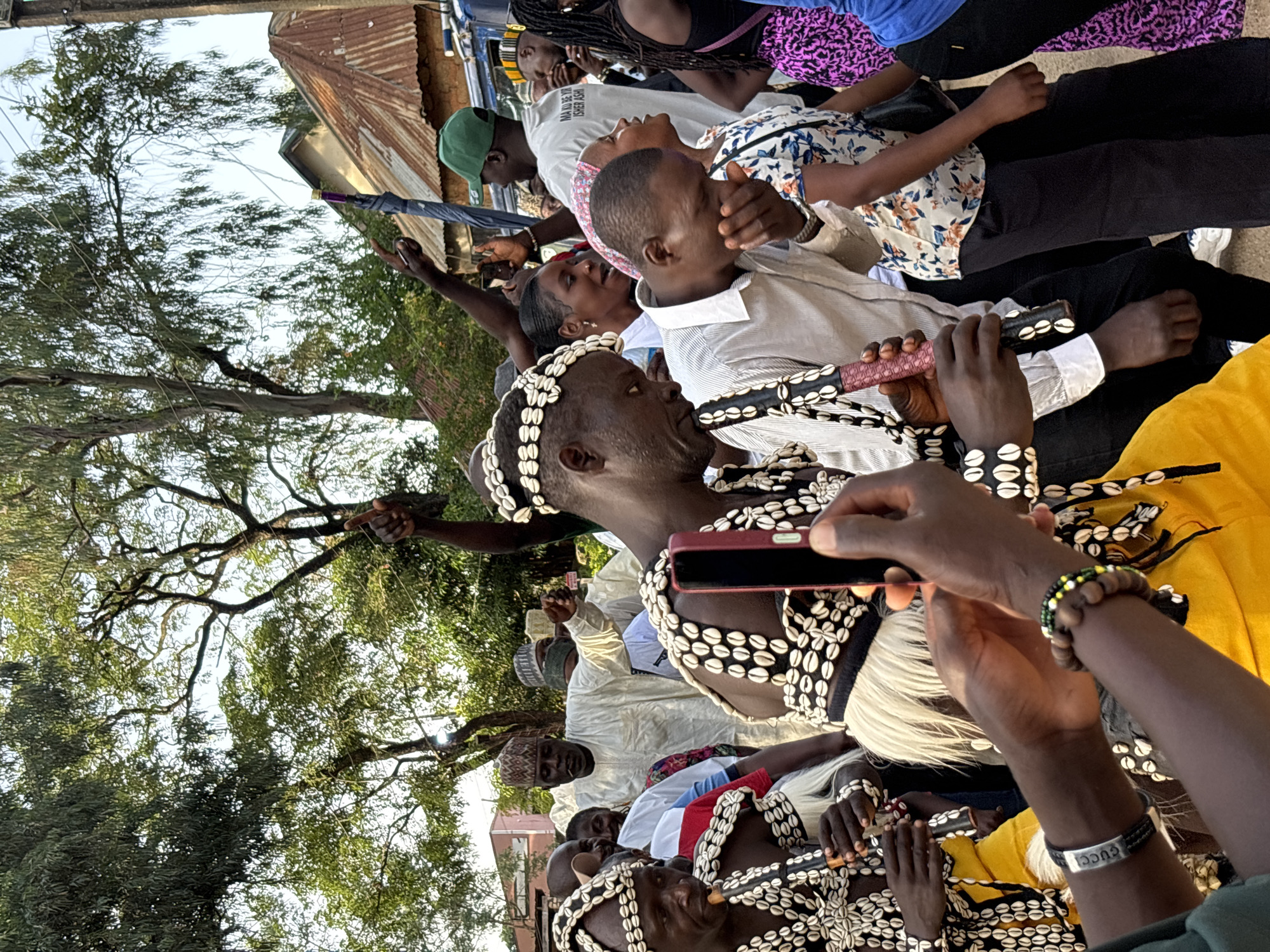 This image depicts a gathering of people in what appears to be an outdoor setting in Jos, Nigeria. The subjects are predominantly men, many adorned in traditional attire. Some are wearing elaborate headdresses or necklaces made of shells. One man in the foreground is playing a traditional instrument, possibly a flute or a horn, which is also decorated with shells. Another individual is holding a smartphone, seemingly taking a picture or recording the event.
The scene is filled with a sense of community and celebration, suggested by the close proximity of the individuals and their engaged expressions. The presence of trees and a rustic corrugated roof in the background indicates a semi-rural or older urban environment. The lighting suggests it is daytime, possibly late afternoon, given the warm tones.
There is visible text on a white t-shirt worn by one of the individuals, which reads: "NA NO BE ISHER ASHI". The overall atmosphere is one of cultural festivity and communal gathering.