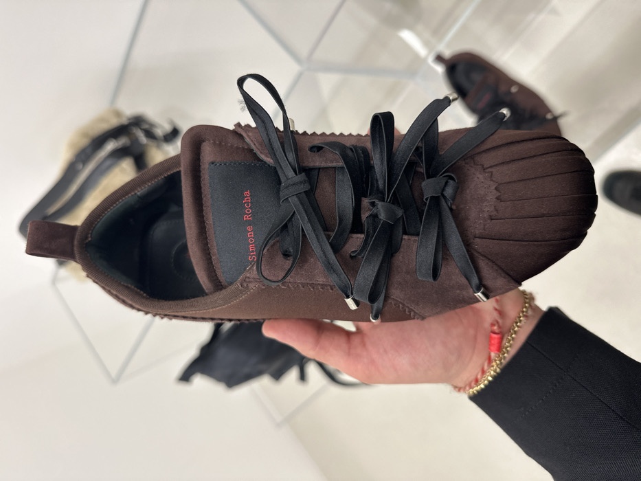 The image displays a close-up of a person's left hand holding a single dark brown Simone Rocha brand sneaker. The shoe features a pleated toe box, a textile upper with visible stitching and texture, and black ribbon-style laces with silver-colored aglets, tied into several small bows. A black label on the shoe's tongue presents the brand name "Simone Rocha" in red capital letters. The person's wrist is adorned with two bracelets: a thin gold-toned chain and a red string bracelet with a small red accent. A black garment sleeve is visible on the forearm. The background is a brightly lit, minimalist retail environment with predominant white or light-colored surfaces and clear acrylic or glass display shelving. Blurry objects in the background include a light-colored, possibly fur or shearling-trimmed, handbag or accessory on an upper shelf to the right, and other dark, indistinct items on lower shelves and the floor. This scene is located within a Simone Rocha retail establishment, specifically identifiable as potentially the Simone Rocha boutique at 31 Rue des Archives in Paris, France.