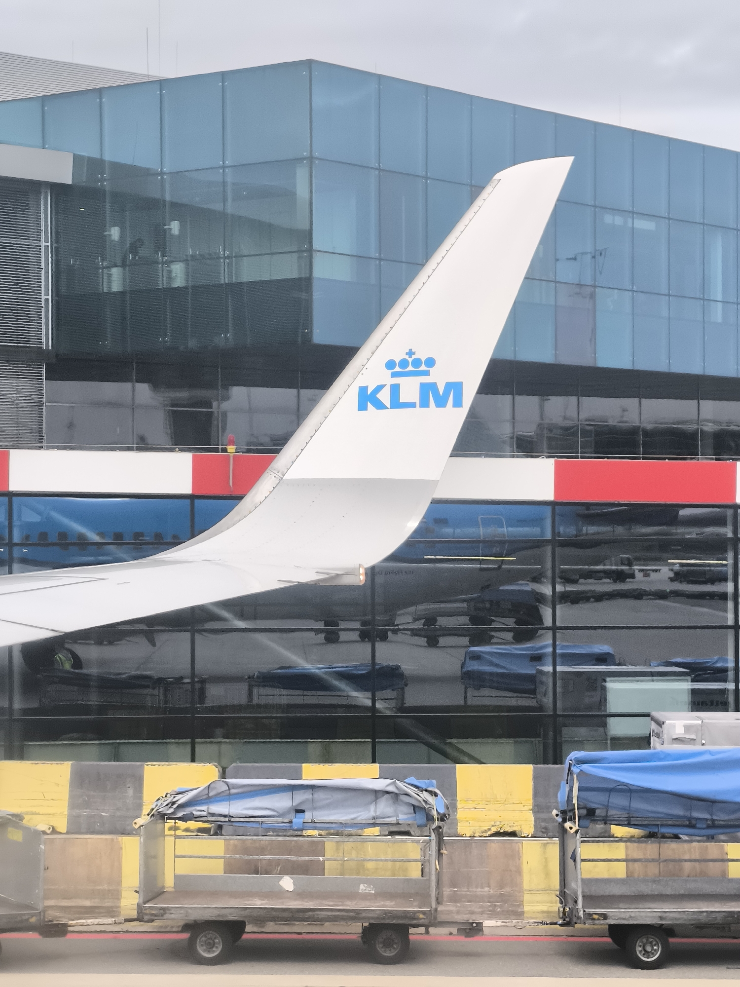 The image shows the tail fin of a KLM airplane with the KLM logo prominently displayed. The tail fin is white with the blue KLM lettering and crown symbol. The background consists of a modern airport building with large glass windows reflecting the surrounding environment. Below the building and partially obscuring the reflection are cargo carts with covered items, likely luggage or freight. The carts have yellow and black striped bumpers. The location is likely an airport, but the specific city is unknown. The sky is overcast, suggesting neutral weather conditions.