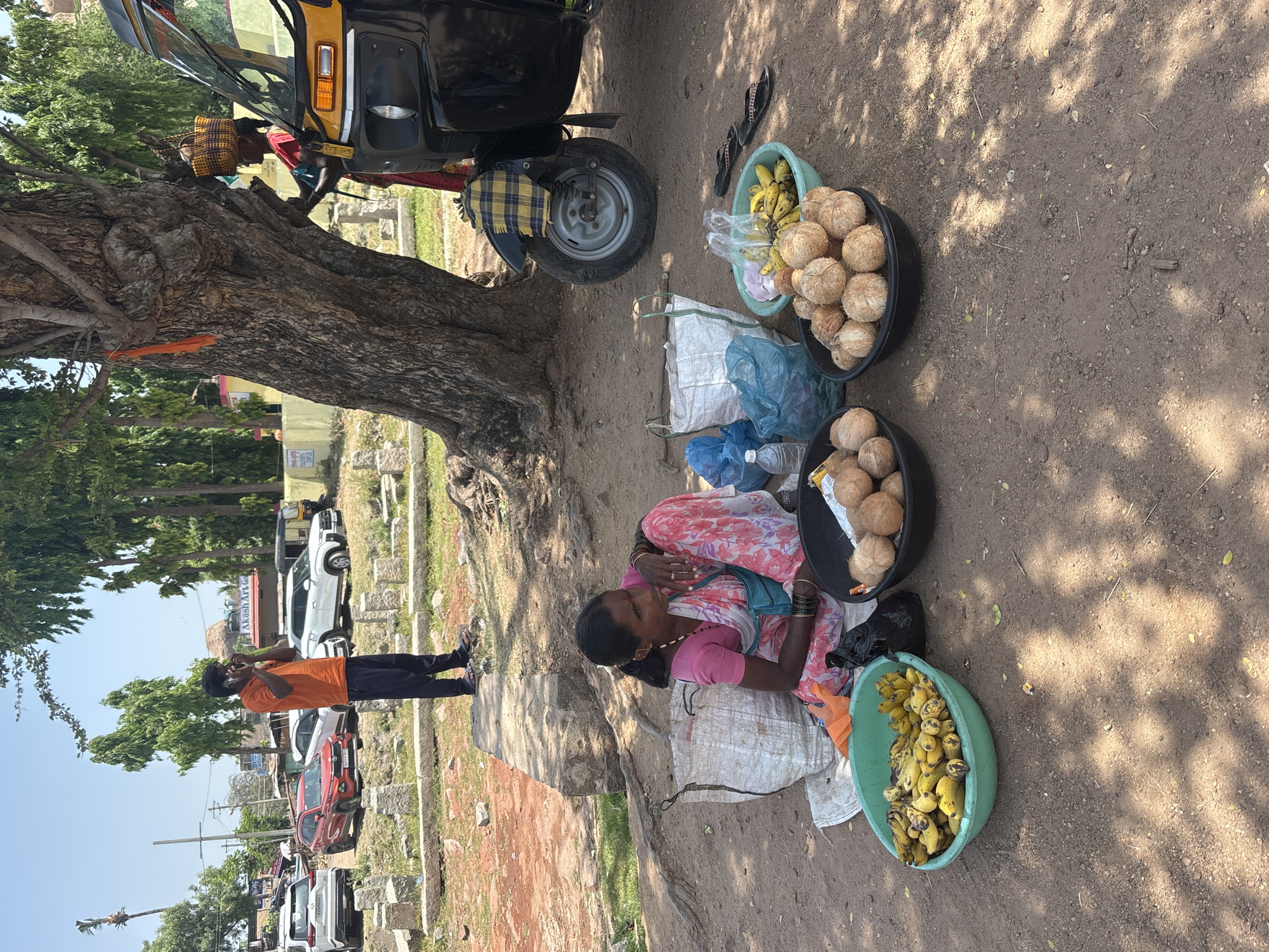 The image depicts a street scene in Kamalapura, India. A woman is sitting on a sack on the ground, surrounded by three large bowls containing coconuts and bananas. These items are likely for sale. Beside her is a parked black auto-rickshaw. In the background, a man in an orange shirt is standing on a street with parked cars. A large tree provides shade over the woman and her merchandise. The ground is dirt and scattered with leaves.