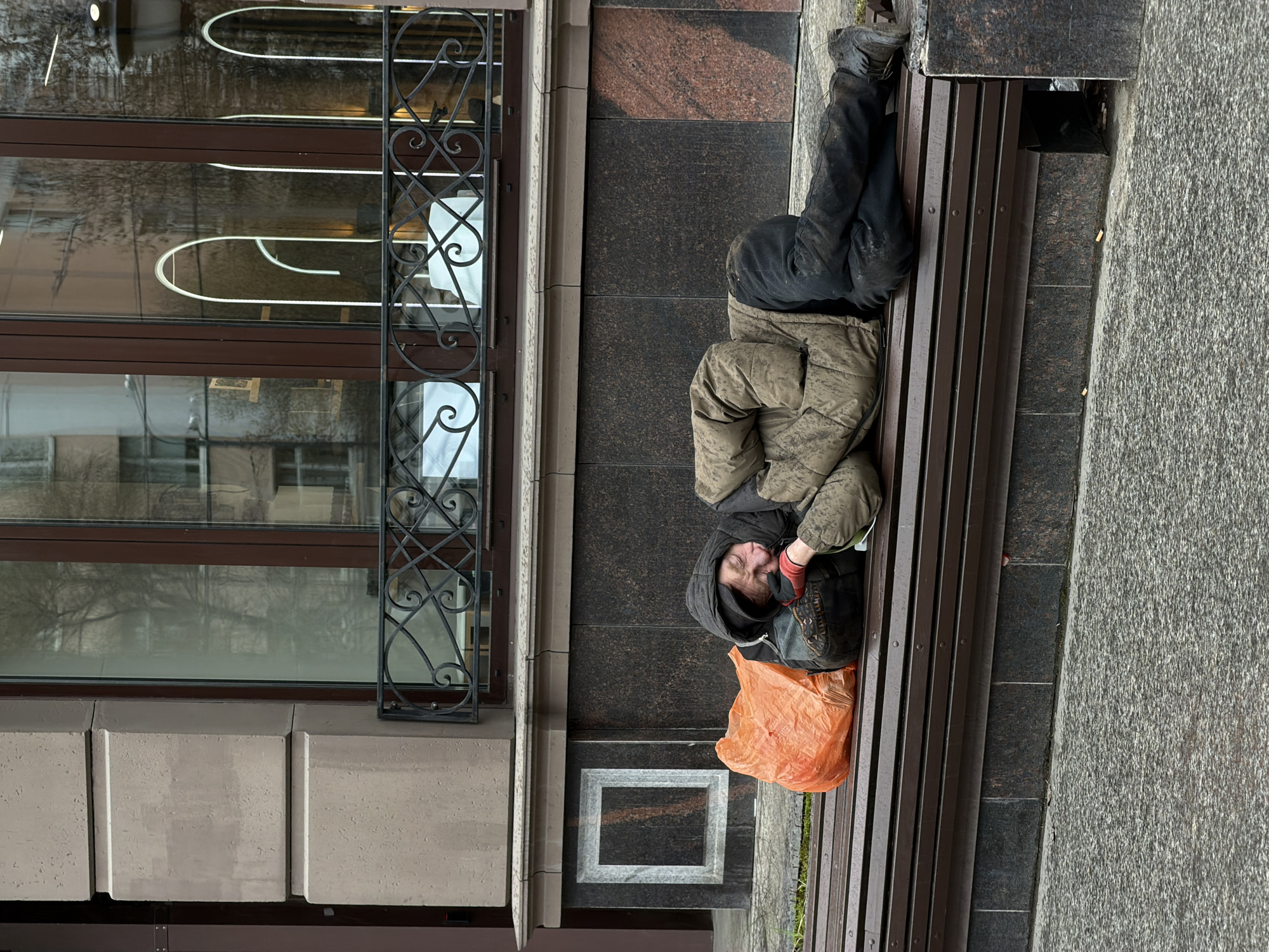 A man is lying on a bench in an urban setting. The man is wearing dark pants, a patterned jacket, and a dark hood. His face is visible, and he is wearing red gloves. An orange plastic bag sits near his head on the bench. The bench is made of dark wood slats. Beside the bench is a sidewalk made of textured grey stone. The bench is situated next to a building with a dark brown tiled wall and a light-colored stone base. A window with ornate black metal scrollwork is visible. The building's architecture appears urban. The location is Unknown, Unknown.
