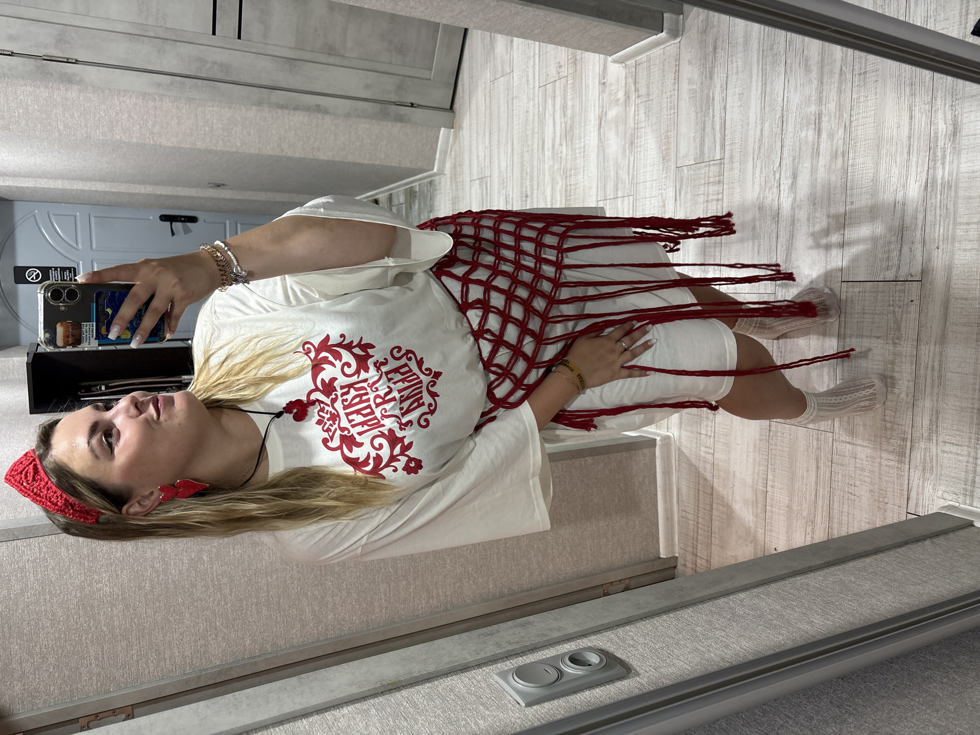 In a domestic setting, a young Caucasian woman with blonde hair and fair skin stands in front of a full-length mirror, taking a selfie with her smartphone. She is wearing a white oversized t-shirt with red embellishments, a red crocheted headband, red heart-shaped earrings, a red necklace with a heart pendant, a red macrame belt with fringe, white socks, and clear/translucent shoes. The background appears to be a hallway or entryway, with textured walls, a wooden door, and a wooden floor. The lighting is artificial and casts shadows. The woman's expression is neutral to slightly smiling, and she is looking towards her reflection. The overall impression is of a casual, self-styled pose within a residential interior. The mention of Kungur, Russia, is not supported by any visual evidence in the image.
