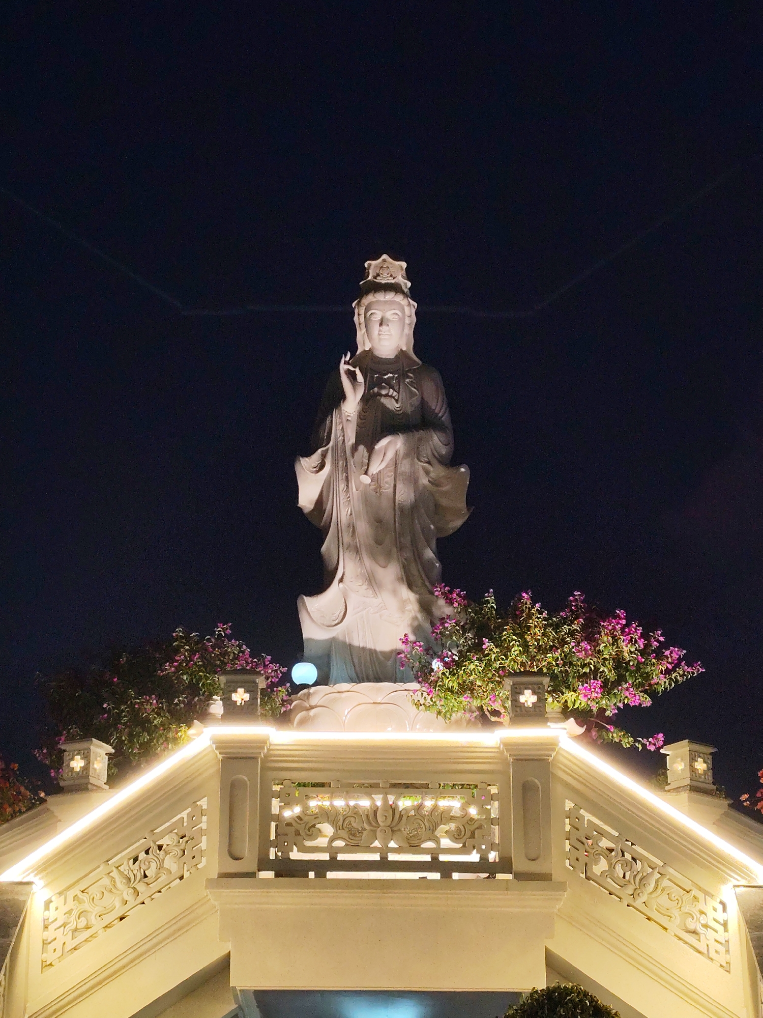 The image shows a statue of a Buddhist figure, likely Quan Yin, in the city of Lâm Viên - Đà Lạt, Vietnam. The statue is made of white stone and stands on a white stone platform with a decorative railing. The railing is illuminated by LED lights. The platform is decorated with flowering plants. The background is a dark night sky. Two faint contrails are visible in the night sky.