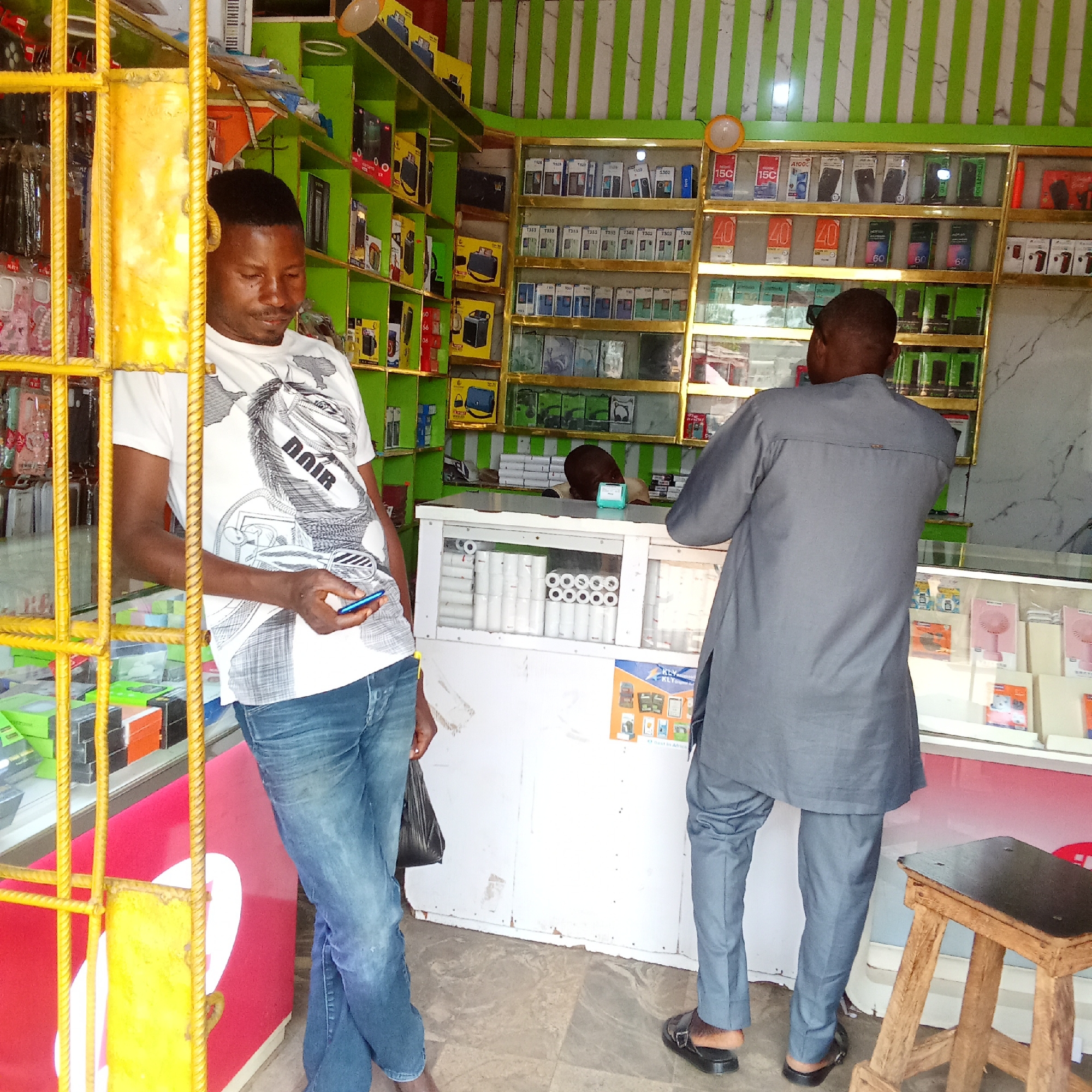 Here is a forensic description of the image:
**Scene:** The interior of a small mobile phone shop, possibly located in an urban or semi-urban area.
**People:**
* A man is standing near the entrance, leaning against what appears to be a metal security gate. He is dressed casually in a white t-shirt with a print, blue jeans, and is holding a mobile phone.
* Another man, wearing a gray shirt and trousers, stands in front of a counter, presumably as a customer.
* A third person, partially visible behind the counter, is likely the shopkeeper.
**Objects:**
* Mobile phones and accessories are displayed on shelves and in glass display cases.
* A counter in the center of the shop contains accessories, such as rolls of paper.
* A stool is positioned near the counter.
* A shopping bag can be seen near the man with the phone.
**Environment:**
* The walls are painted with green stripes.
* The shop is well-lit, and the interior appears to be cluttered with merchandise.
**Actions and Interactions:**
* The man at the entrance is looking at his phone.
* The other man is likely interacting with the shopkeeper regarding a purchase or inquiry.
**Location:** City of Unknown, Unknown. No further geographical context is discernible from the image.