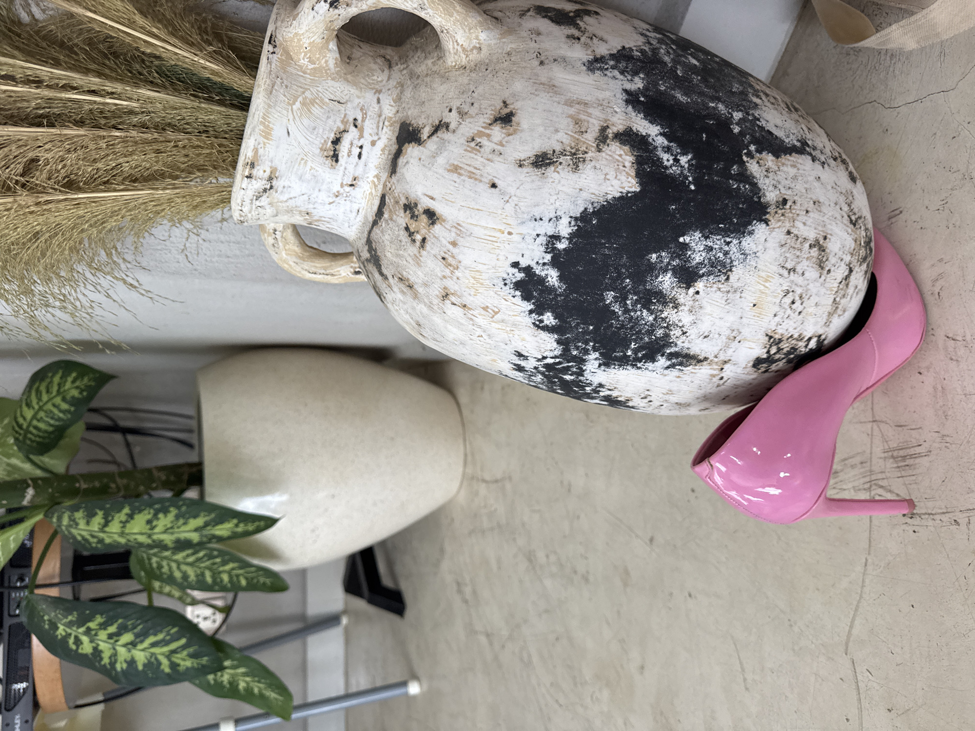 The image shows a still life arrangement on a grey floor. A large, aged white vase with black markings and handles rests on its side, its opening filled with dried pampas grass. A glossy pink high-heeled shoe is positioned near the vase. To the left, partially visible, is a white pot containing a plant with green and yellow patterned leaves. The image suggests an indoor setting, possibly a studio or staging area. Ungasan, Indonesia is not visible or identifiable in this image.