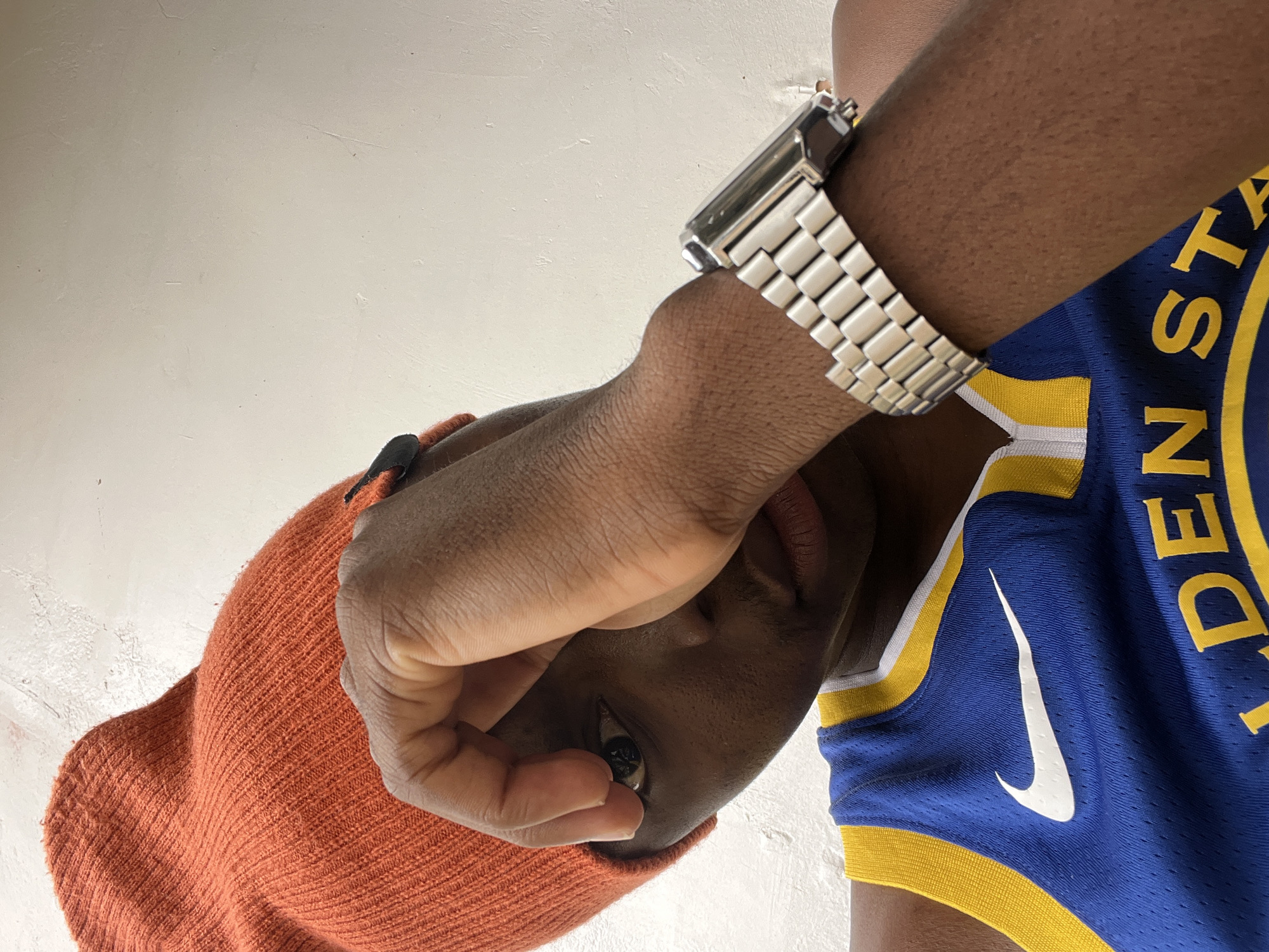 This media file is a close-up, low-angle, portrait-style photograph of a person. The subject is wearing an orange knitted beanie and a blue and yellow basketball jersey with a white Nike swoosh logo. The jersey appears to be a Golden State Warriors jersey, as indicated by visible yellow lettering that reads "DEN ST" from "GOLDEN STATE". The subject also wears a silver metal band watch on their left wrist. Their right hand is partially covering their face, with their thumb and fingers obscuring part of their eye and cheek. The background is a plain, textured white surface, possibly a wall. The lighting suggests it could be daytime, but there are no strong shadows to indicate time of day or specific weather conditions. The setting appears to be indoors, due to the consistent, diffuse lighting and plain background. The overall mood is casual and introspective, with the subject's gaze directed downwards.