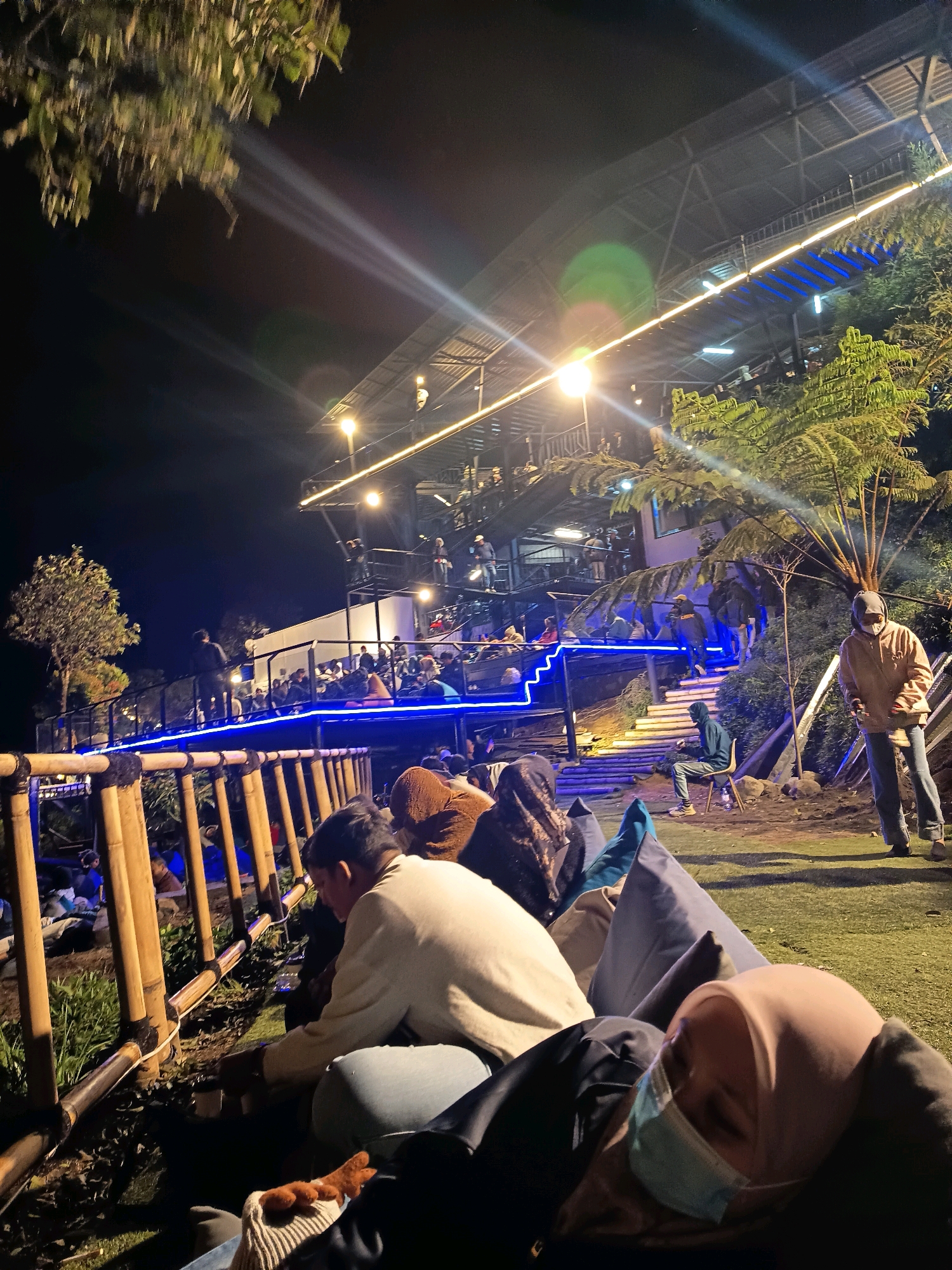 The image depicts an outdoor seating area at night, likely a restaurant or cafe, with multiple levels and a vibrant atmosphere. The location is identified as Keditan, Indonesia.
In the foreground, several people are seated on beanbag chairs on a grassy area. One person in a light-colored sweater and jeans is hunched over, looking down. Another person, wearing a light pink hijab and a face mask, is reclining on a beanbag, seemingly asleep. Their face is partially obscured by their hijab and mask.
Behind the foreground seating area, a bamboo fence separates the grassy space from a raised platform. This platform is illuminated by blue and yellow lights, and a multitude of people are seated at tables, dining and socializing. A set of stairs with blue lights leads up to another level of the establishment, where more people are visible.
Further up, a large, multi-story structure with a metal roof is illuminated by various lights. People are seen standing and walking on different levels of this structure. Tall, leafy trees and ferns surround the area, adding to the natural ambiance. The overall scene suggests a lively gathering, possibly a tourist destination given the multiple levels and open-air setup. A person in a tan hoodie and jeans stands on the right side of the image, looking towards the seating area.
