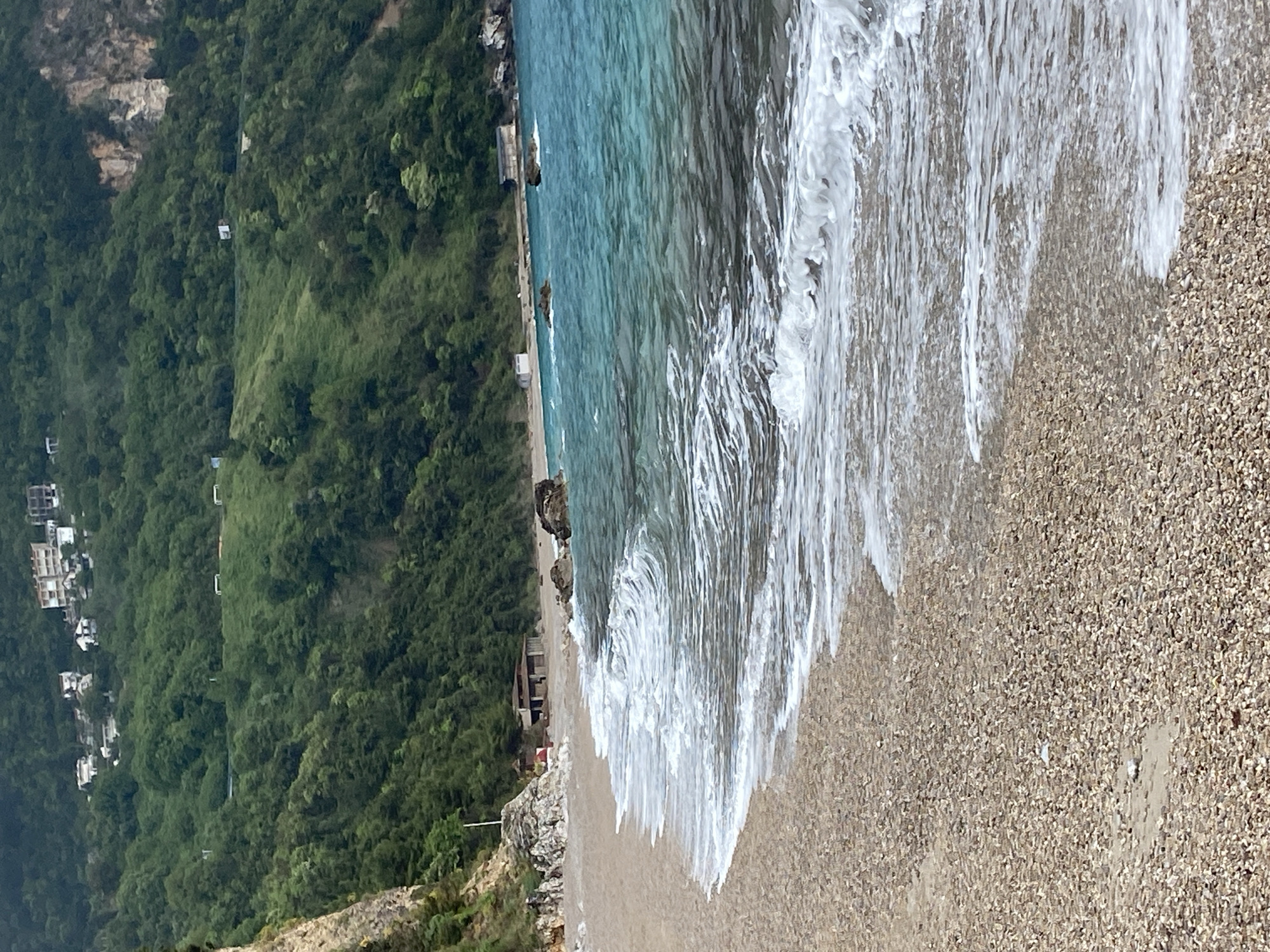 This image presents a serene coastal landscape in Prijevor, Montenegro, featuring a beach, the sea, and a verdant mountain.
On the right side of the frame, the clear, turquoise-blue sea meets the shore. Gentle waves are actively breaking, creating extensive patterns of white foam that spread across the light brown, pebbled beach. The water appears shallow and translucent near the shore, allowing some visibility to the seabed.
The central section is dominated by the pebble or fine gravel beach, stretching horizontally across the view. The beach itself is devoid of people, contributing to a tranquil atmosphere. Along the base of the mountain, where it meets the beach, a few small, simple structures, possibly beach kiosks or changing rooms, are visible.
To the left, a steep, densely forested mountain rises. It is covered in lush green trees and thick vegetation. Higher up the mountain slope, towards the left, a small cluster of white buildings, likely residential, is nestled among the trees. A faint, dark line, possibly a cable car or utility line, can be observed traversing the mountainside, with several small, white support structures attached.
The scene is illuminated by bright daylight, suggesting a clear and sunny day. The overall impression is one of natural beauty and peacefulness in a picturesque Adriatic setting. No visible text is present in the image.