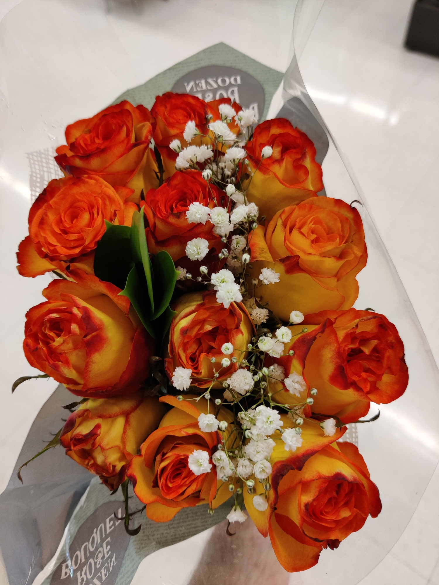 A bouquet of orange and yellow roses, accented with small white flowers, is presented in a clear plastic wrap. The roses are arranged in a cascading fashion. The background is a plain white surface. There is text in a circular gray label that is difficult to read. No people are visible in the image.