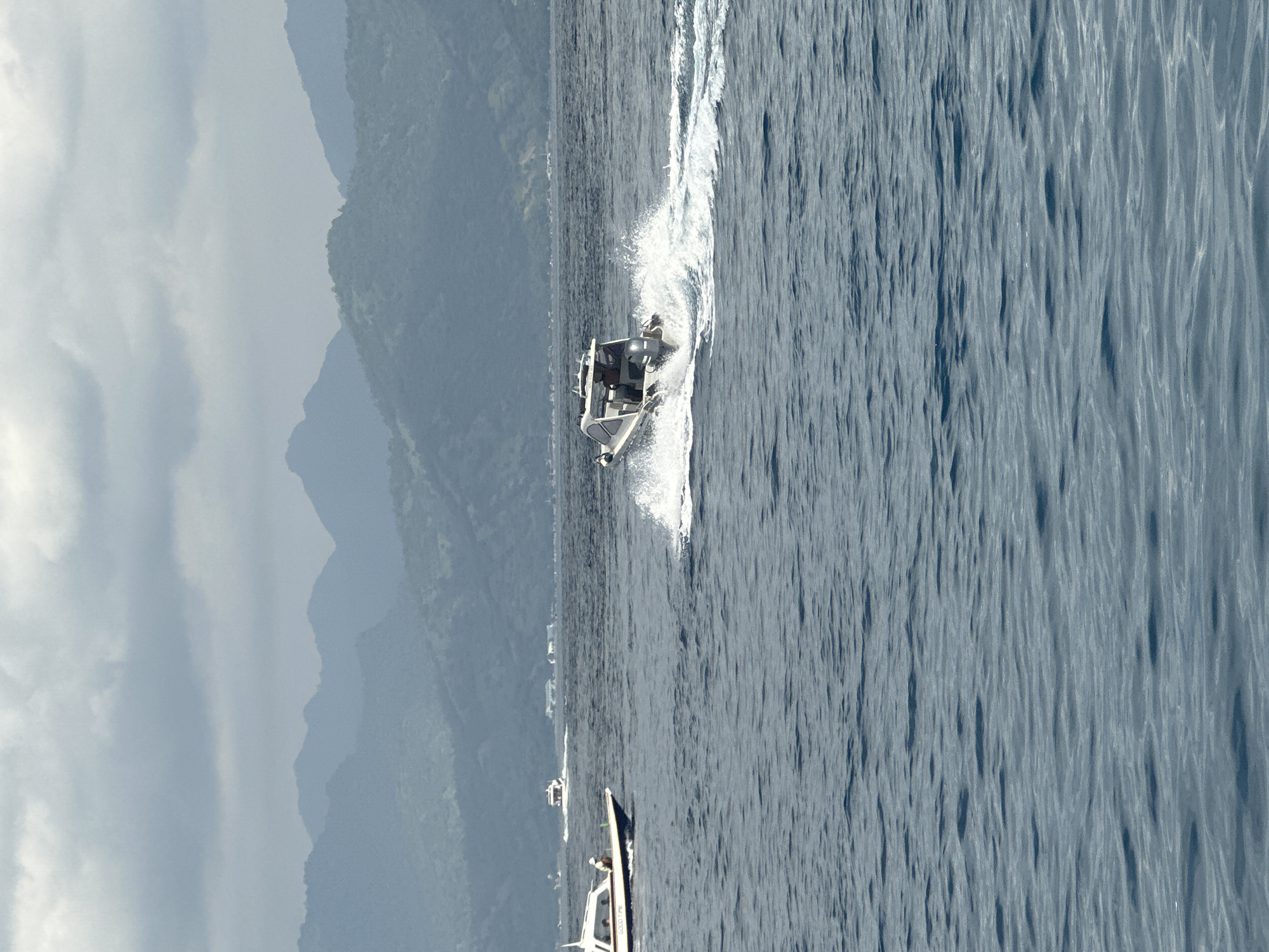 The image shows a white boat moving across the ocean, creating a white wake. In the background, there are mountainous land masses. The location is in Unknown, Indonesia. There are two other smaller boats in the distance. The sky is overcast. The water is a dark blue. There appears to be one person onboard the boat.